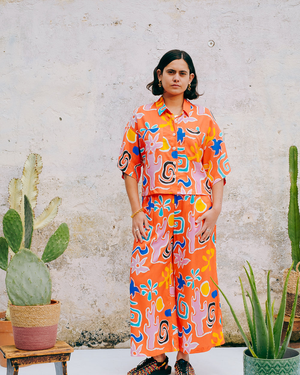 A pair of wide leg cropped trousers with a desert print in vibrant bright orange, featuring front pleats, an elasticated back waist, button and zip on the side, and side pockets.