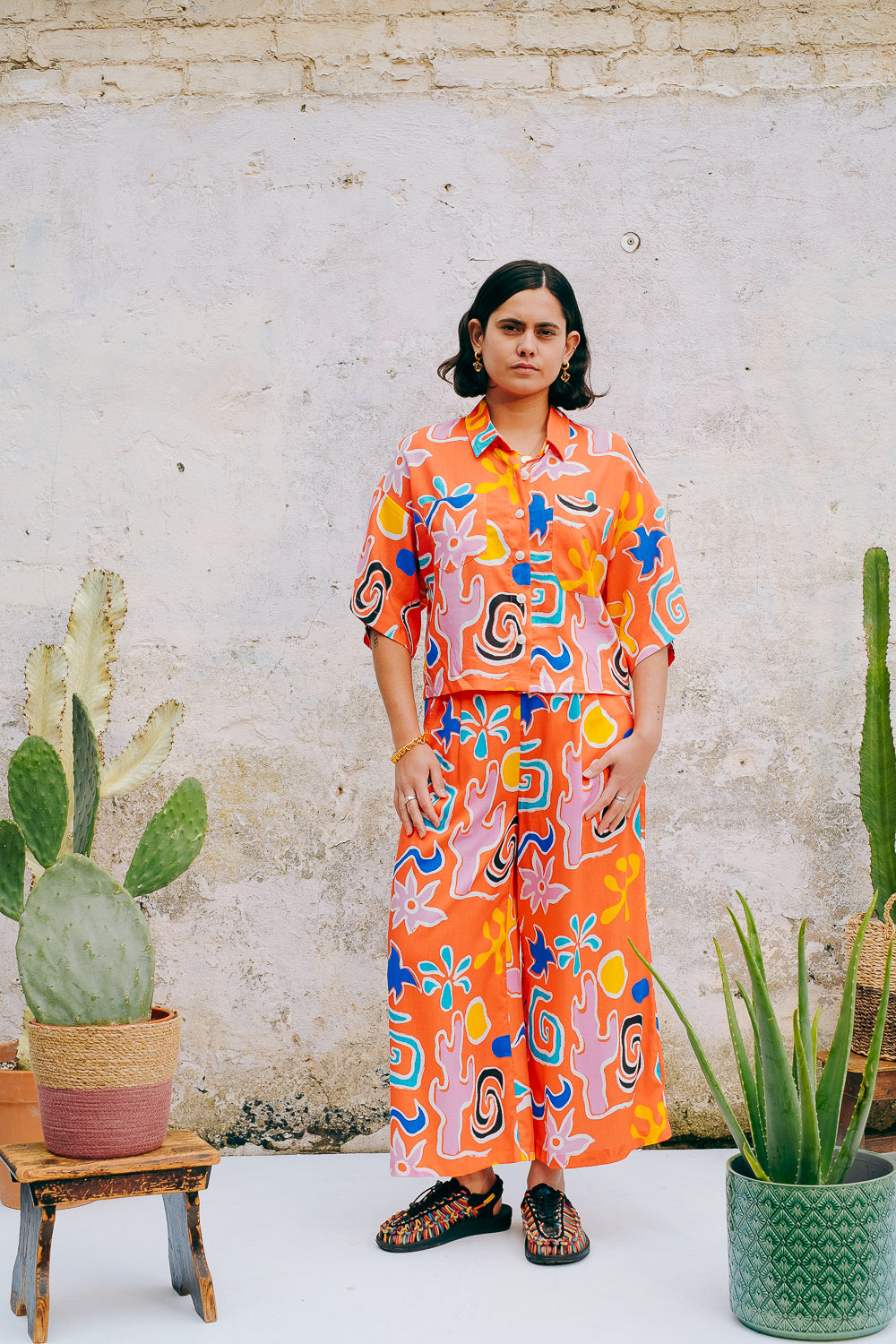 A pair of wide leg cropped trousers with a desert print in vibrant bright orange, featuring front pleats, an elasticated back waist, button and zip on the side, and side pockets.