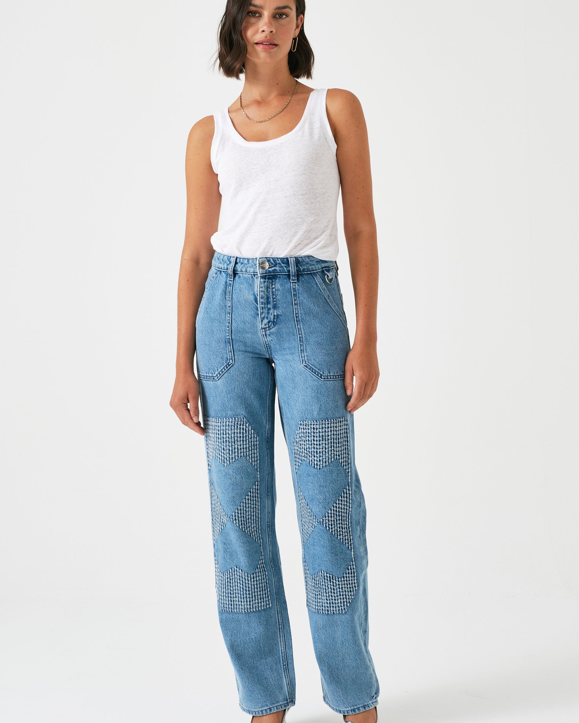 A pair of blue carpenter style jeans with embroidery on the front legs and a center front seam, displayed against a white background.