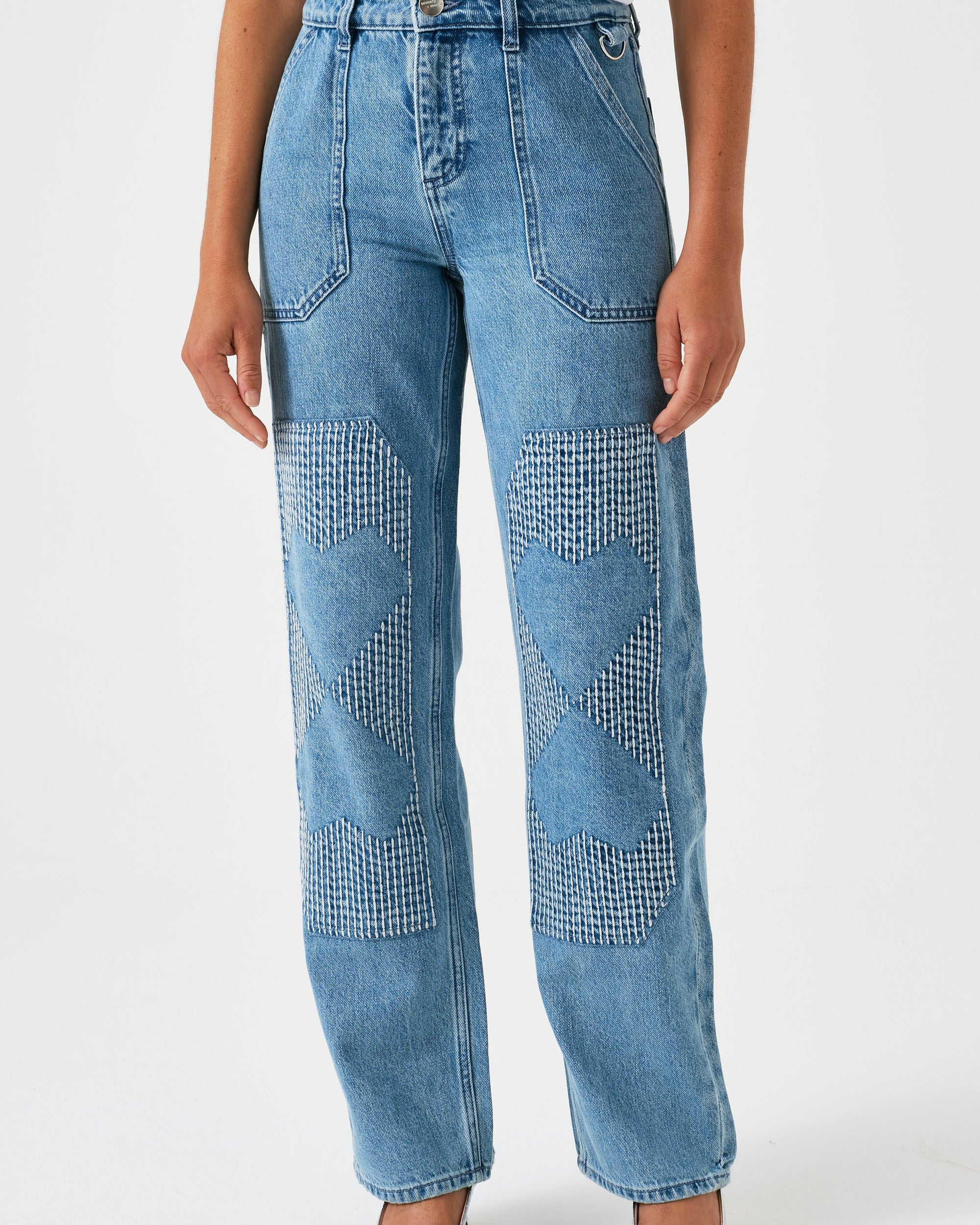 A pair of blue carpenter style jeans with embroidery on the front legs and a center front seam, displayed against a white background.