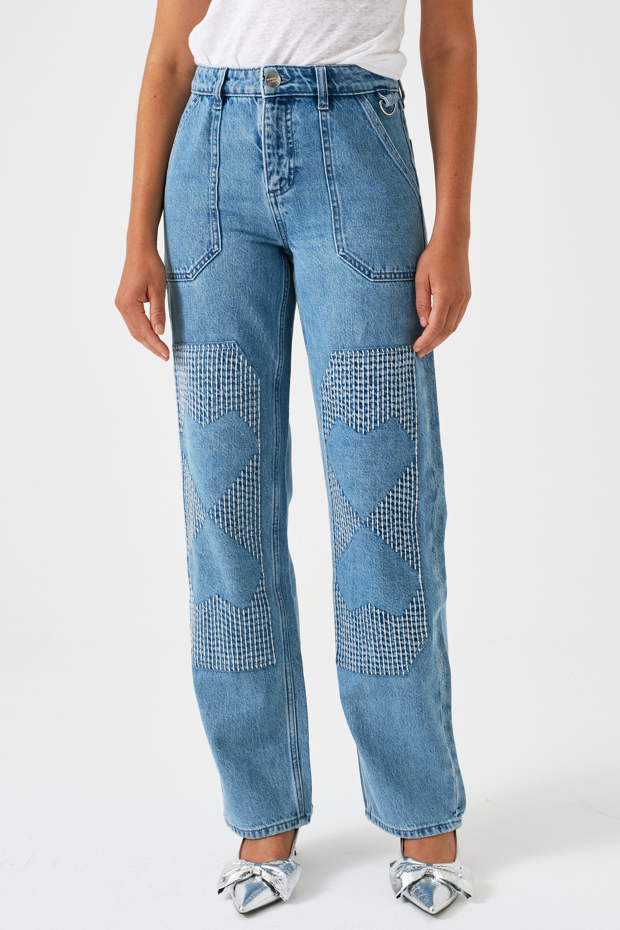A pair of blue carpenter style jeans with embroidery on the front legs and a center front seam, displayed against a white background.