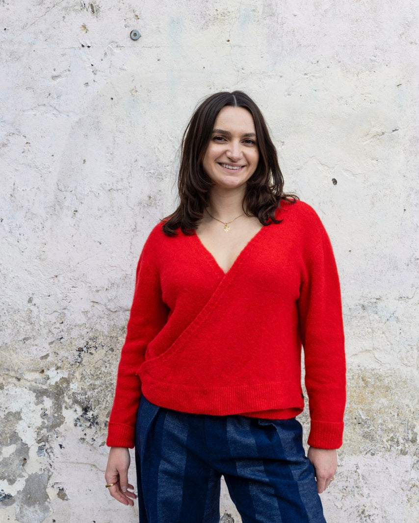 Woman wearing a red sweater and blue pants standing against a textured white wall.