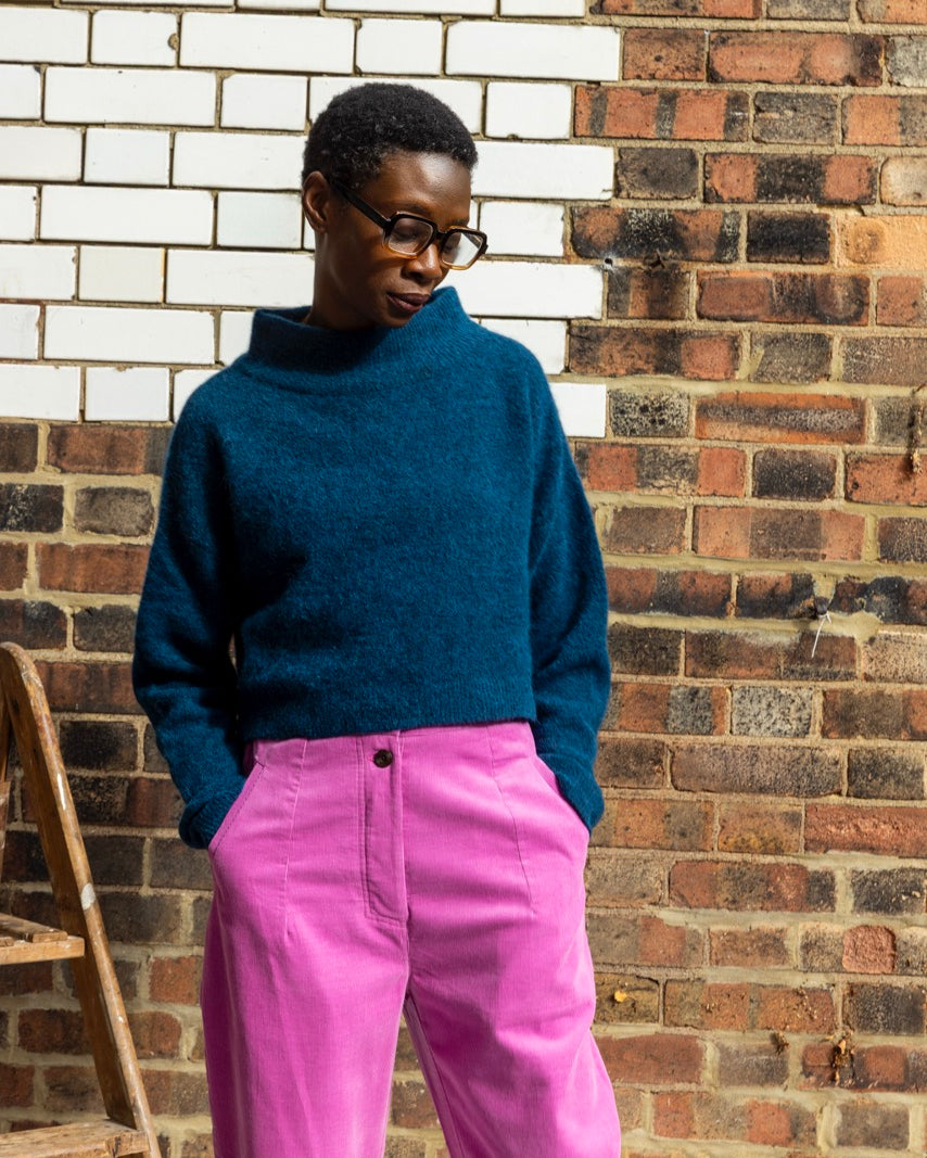 Person wearing a blue sweater and pink pants standing against a brick wall.