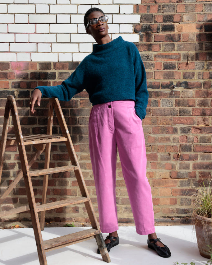 Person wearing a blue sweater and pink pants standing next to a wooden ladder against a brick wall.