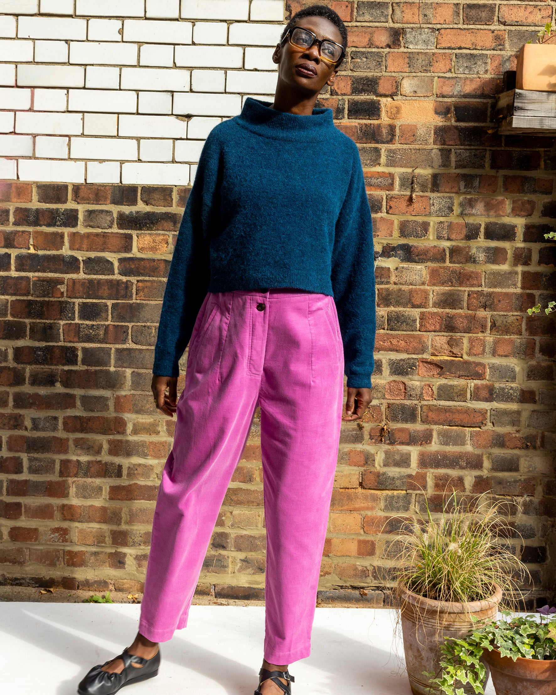 Person wearing a blue sweater and pink pants standing against a brick wall.