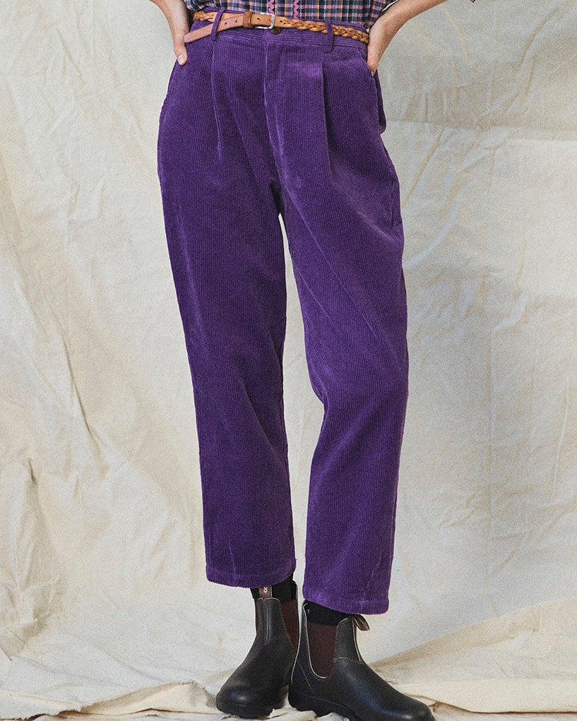 Person wearing purple corduroy pants with a checkered shirt and black boots on a textured beige background