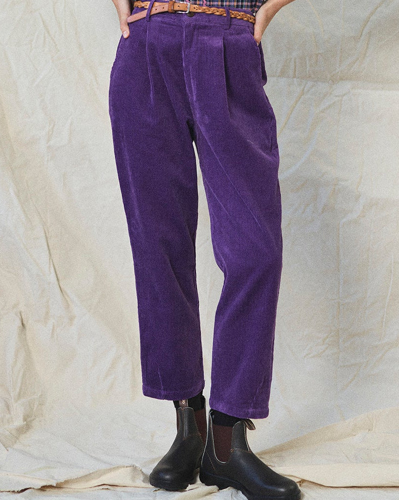 Person wearing purple corduroy pants with a checkered shirt and black boots on a textured beige background