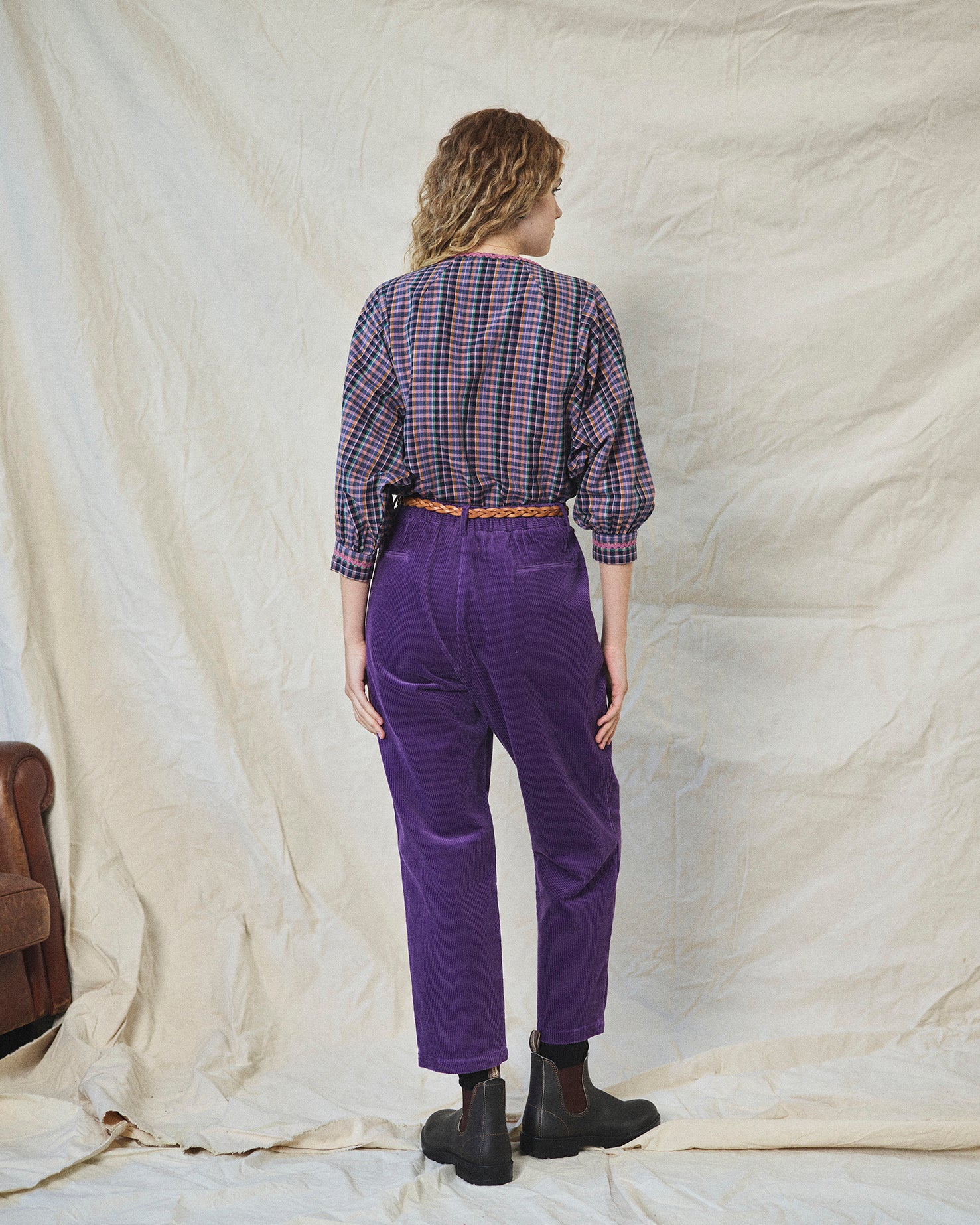 Person wearing purple corduroy pants with a checkered shirt and black boots on a textured beige background