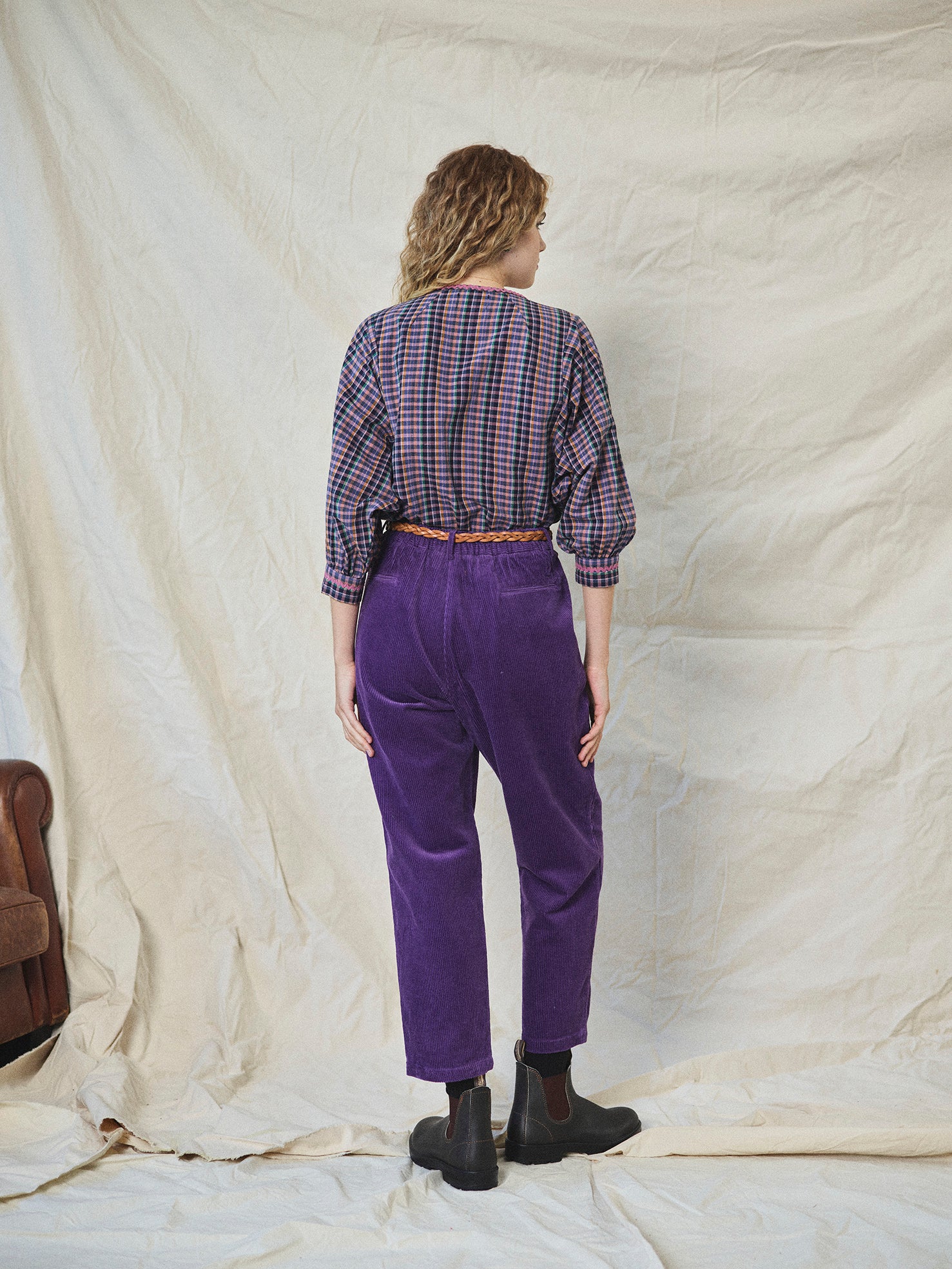 Person wearing purple corduroy pants with a checkered shirt and black boots on a textured beige background