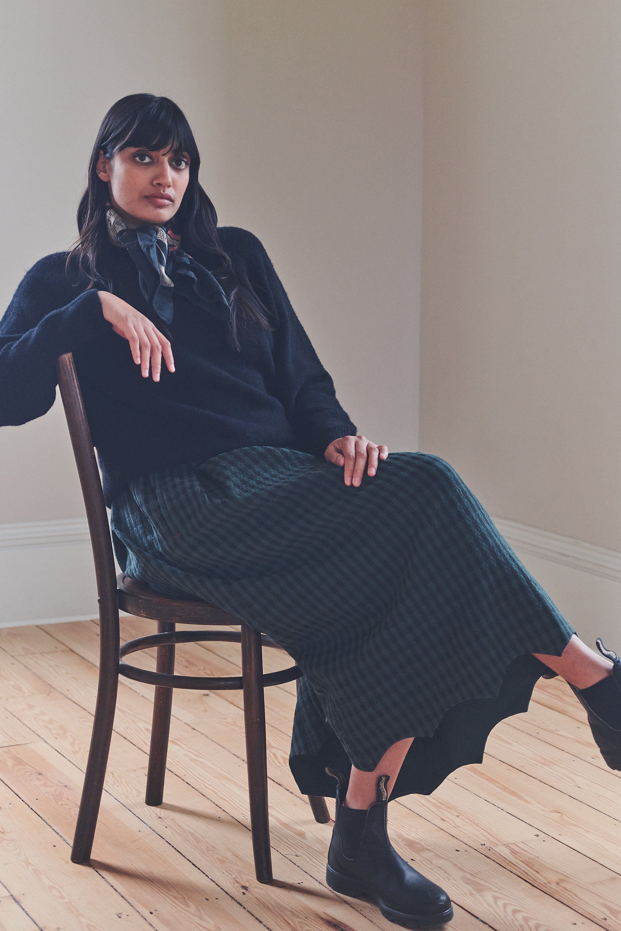 Woman sitting on a chair wearing a dark blue sweater and patterned skirt in a minimalistic room.