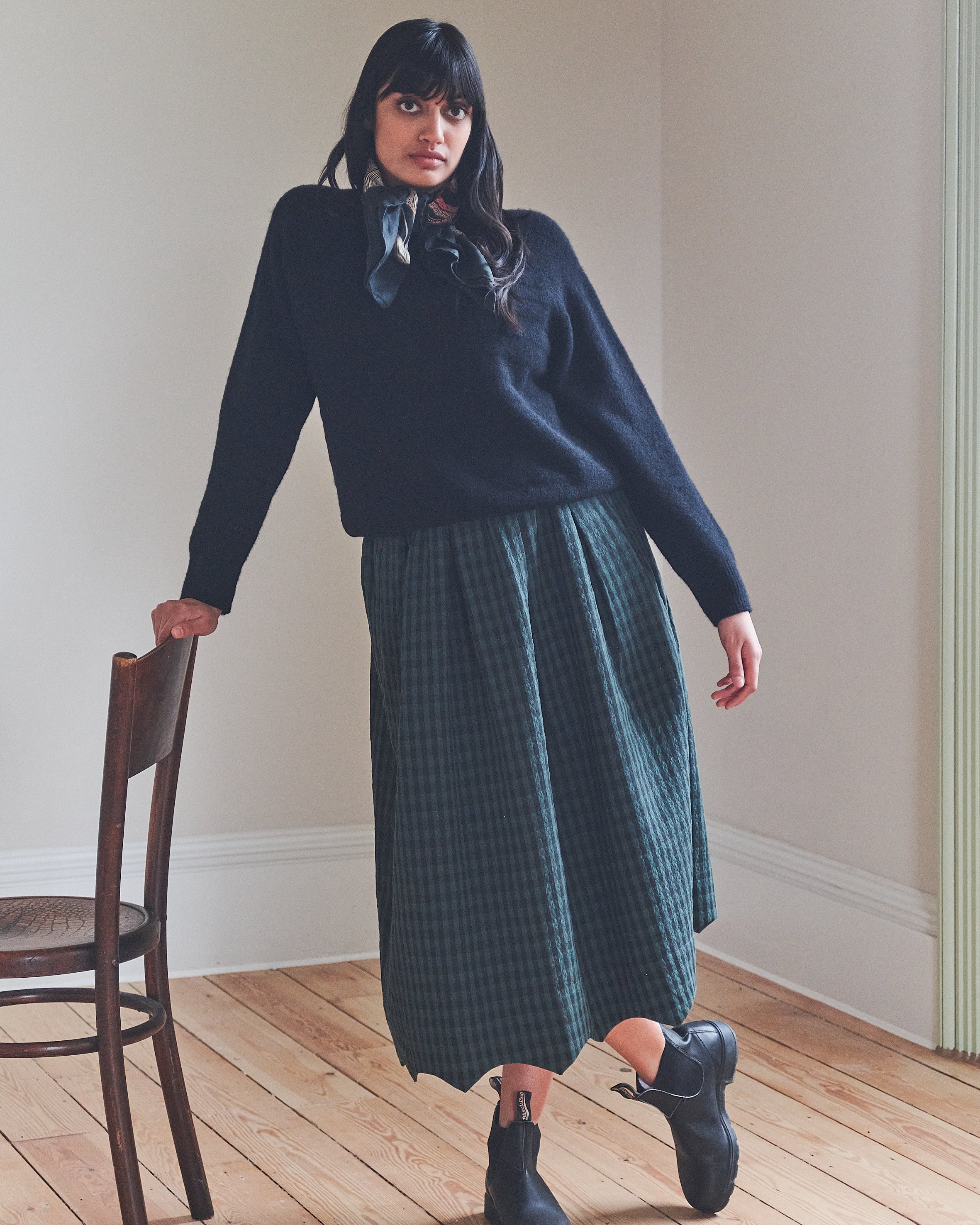 Woman wearing a navy sweater and green plaid skirt standing in a room with wooden floor and white walls.