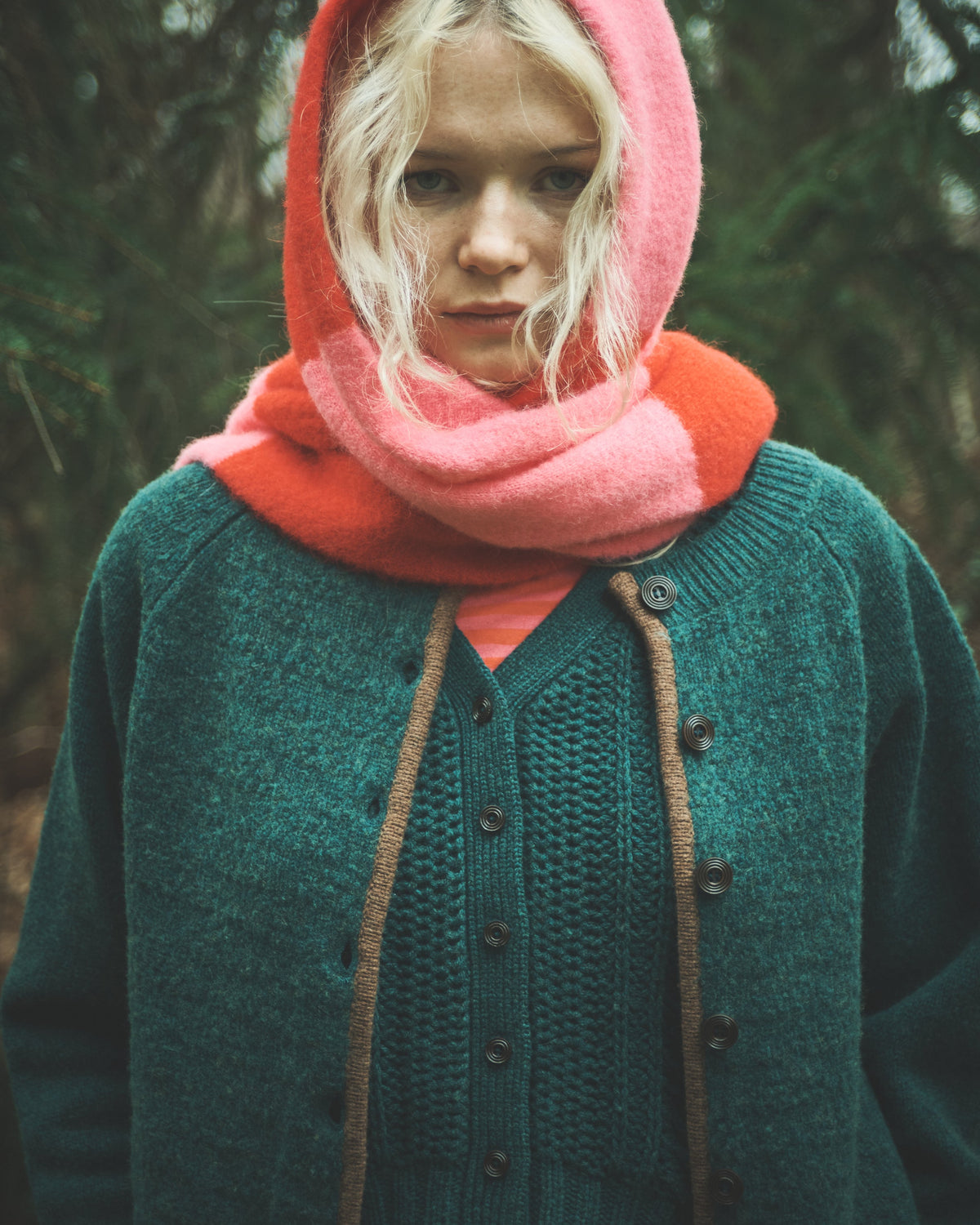 A woman wearing a teal knit cardigan, a bright pink hood, and a coral-orange neck warmer, standing outdoors in a dark, blurred forest setting.
