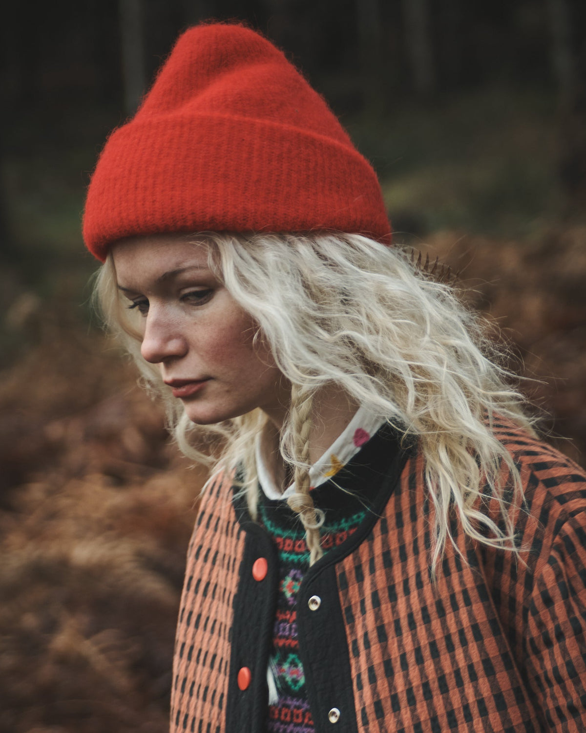 A woman wearing a bright red knit beanie and an orange-and-black checkered jacket, standing outdoors in a dark, wooded setting.