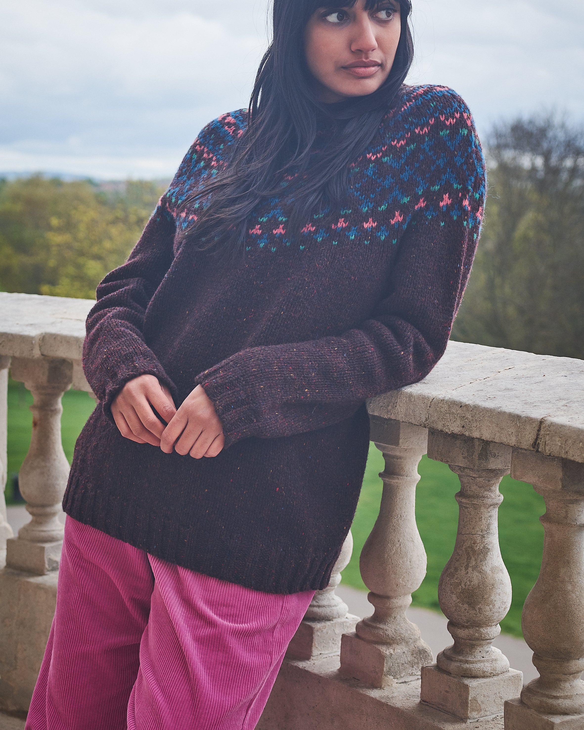 Woman wearing a dark sweater with colorful patterns and pink pants standing on a stone balcony.