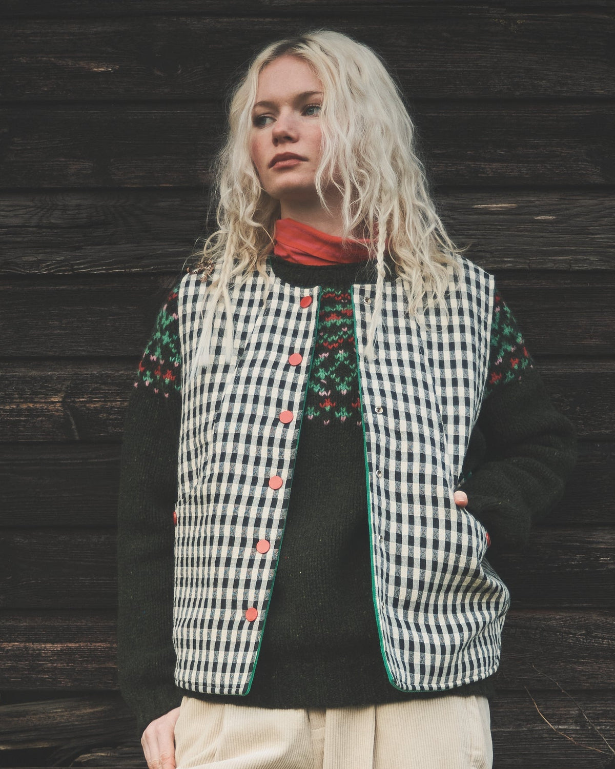 A woman stands outdoors against a dark wooden wall wearing a black and white gingham vest with colorful embroidered details, a black long-sleeve top, and cream-colored trousers.