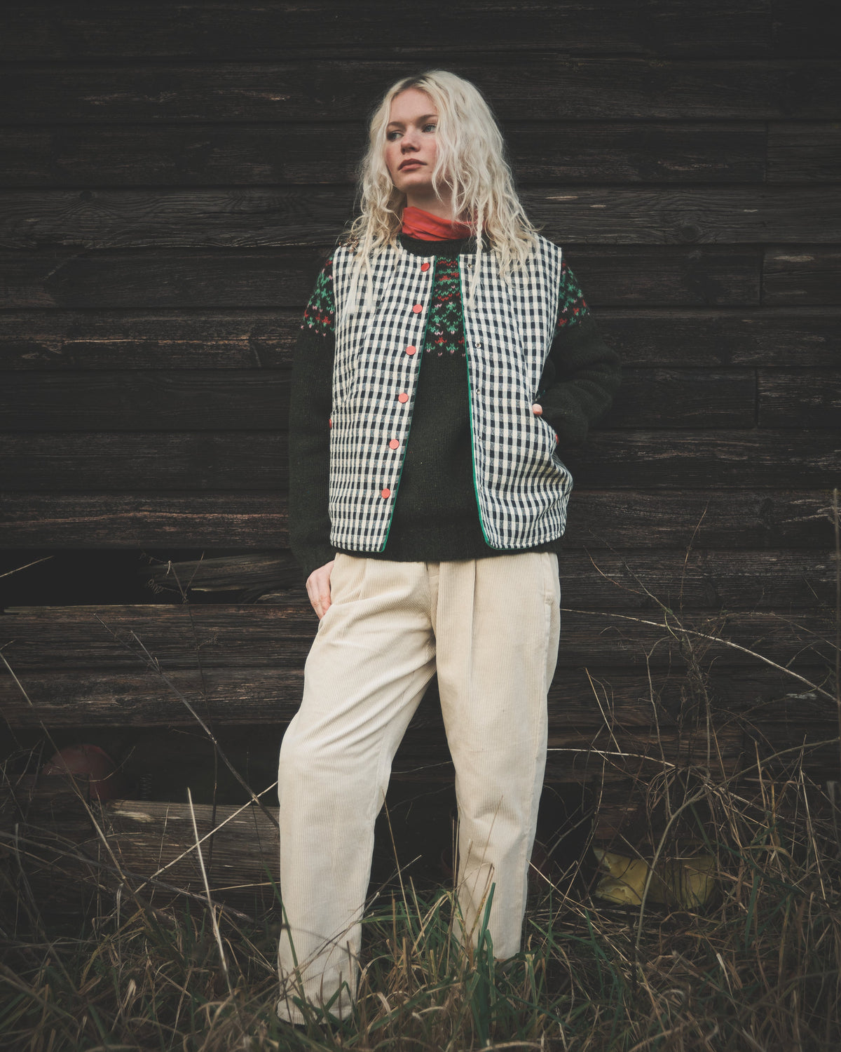 A woman stands outdoors against a dark wooden wall wearing a black and white gingham vest with colorful embroidered details, a black long-sleeve top, and cream-colored trousers.