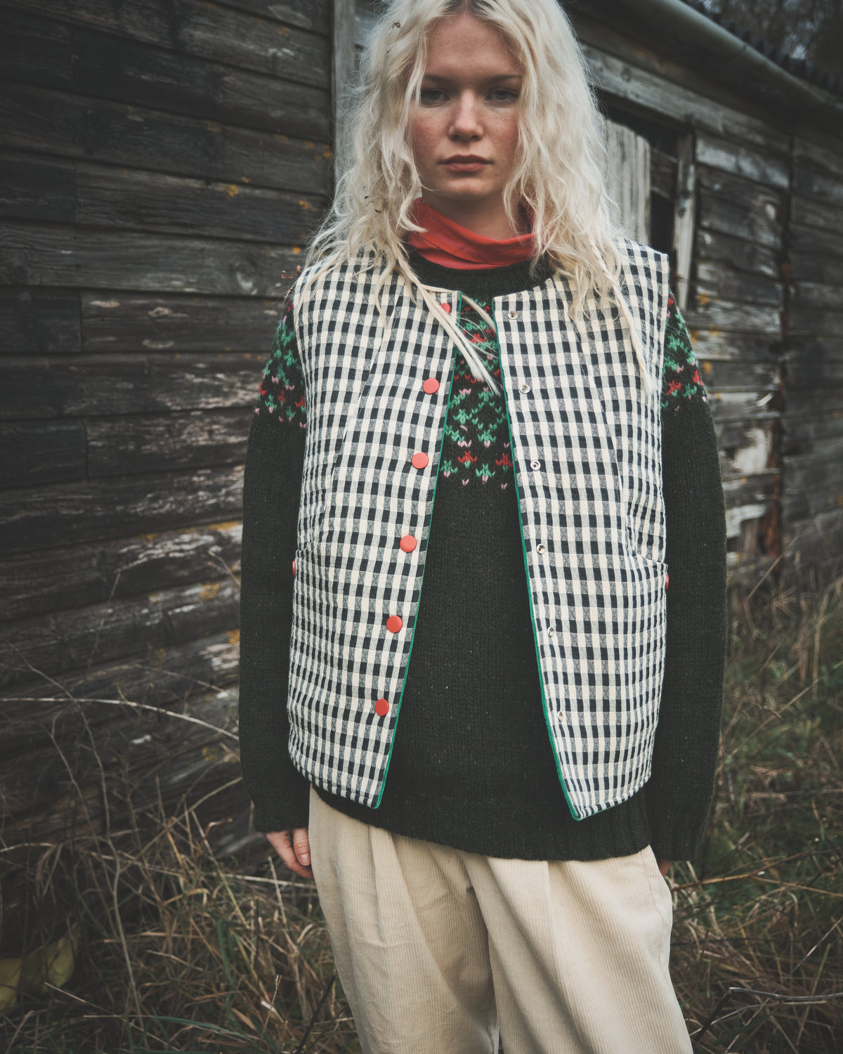 A woman stands outdoors in front of a weathered wooden wall, wearing a black-and-white gingham vest with red buttons and embroidered green floral details on the chest, layered over a dark sweater and light beige pants.