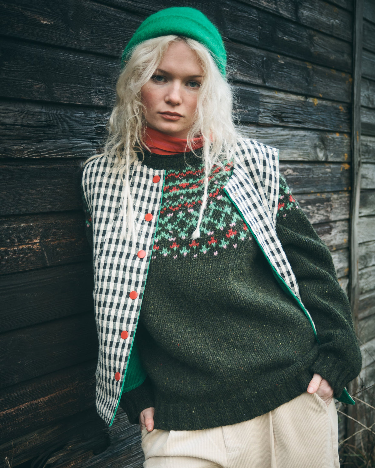 A model wearing a festive Christmas-themed knit outfit, featuring a dark green crewneck sweater with a colorful Fair Isle yoke, a black and white gingham vest with green trim and red buttons, a green beanie, and beige trousers, posed against a dark rustic wooden wall.