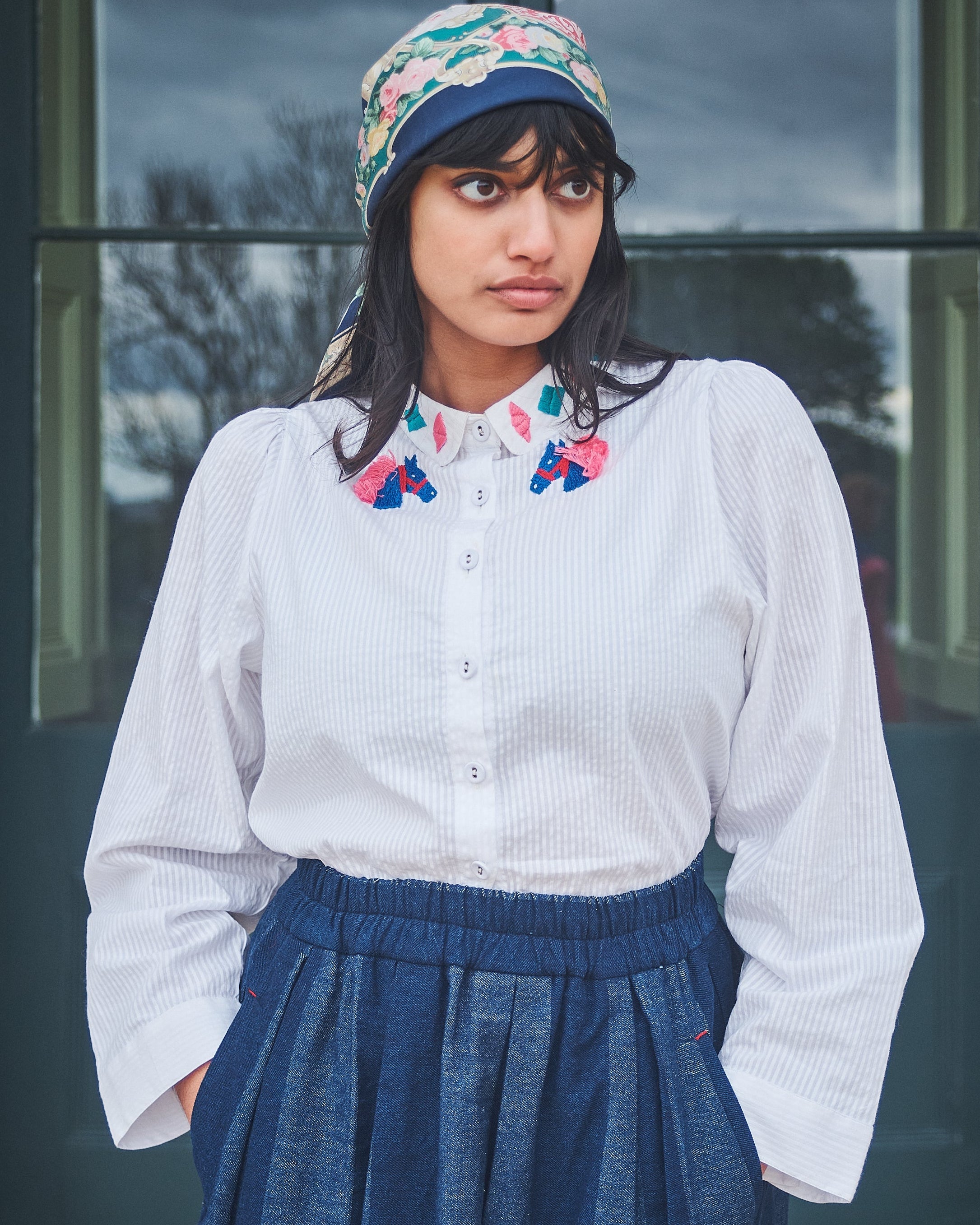 Woman wearing a white blouse with floral collar, blue skirt, and colorful headscarf outdoors.