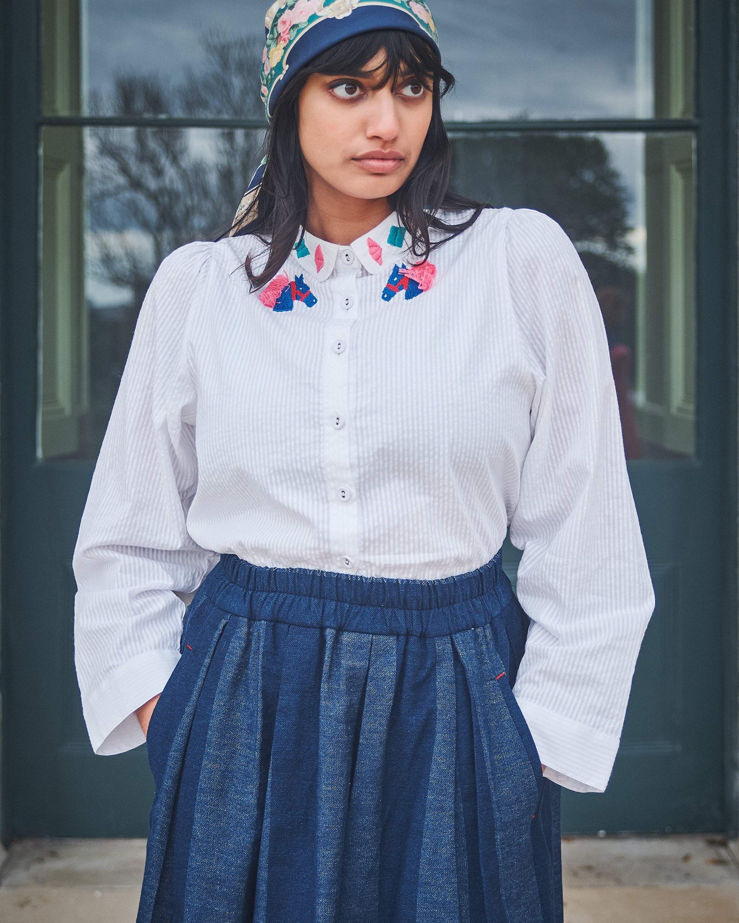 Woman wearing a white blouse with floral collar, blue skirt, and colorful headscarf outdoors.