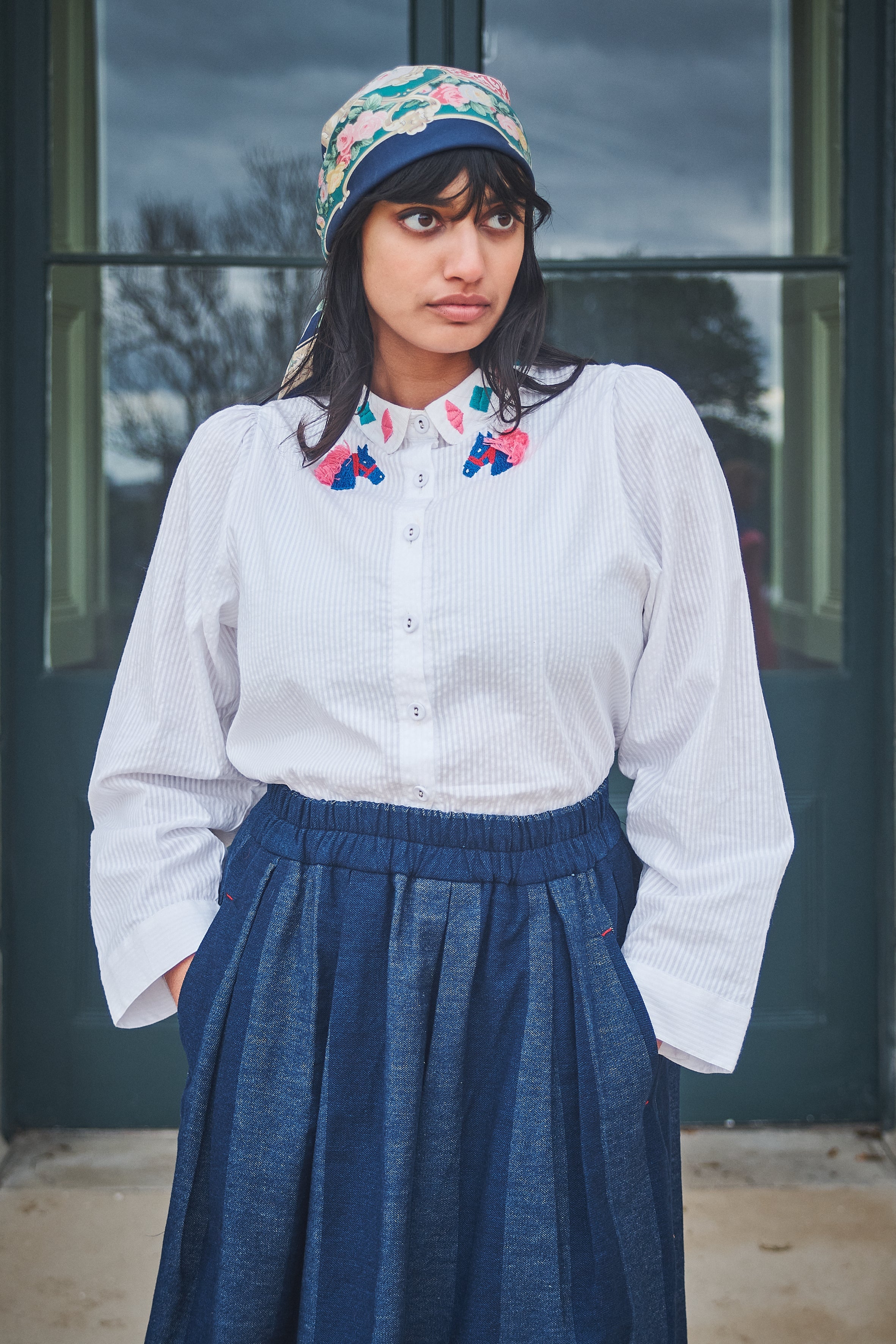 Woman wearing a white blouse with floral collar, blue skirt, and colorful headscarf outdoors.