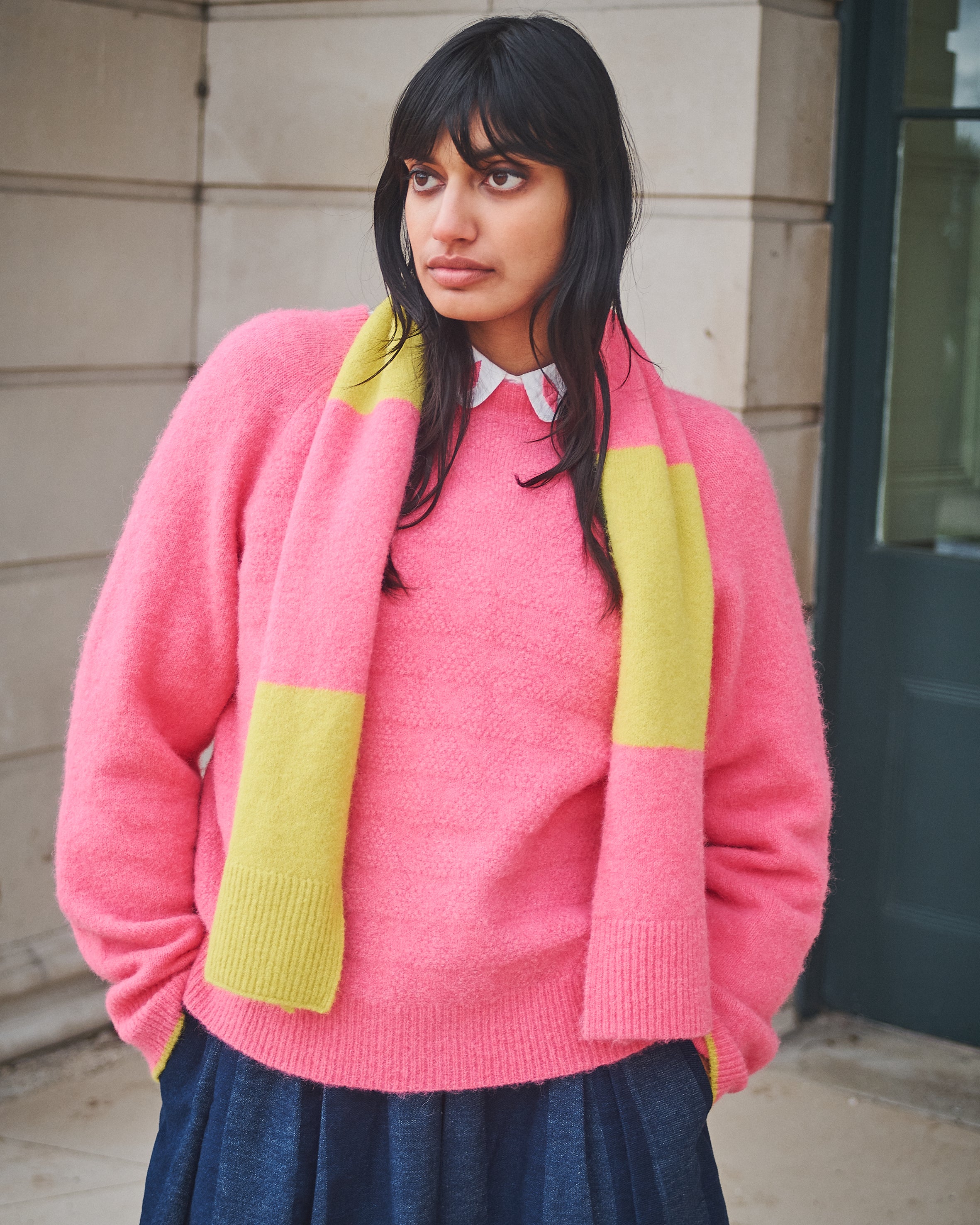 Woman wearing a pink sweater with yellow accents standing against a building.