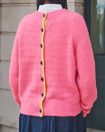 Person wearing a pink cardigan with a yellow trim and black buttons, standing against a neutral background.