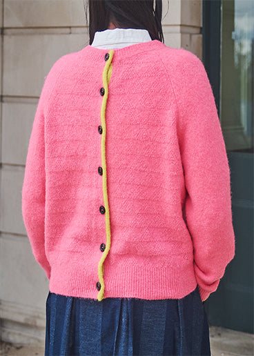 Person wearing a pink cardigan with a yellow trim and black buttons, standing against a neutral background.