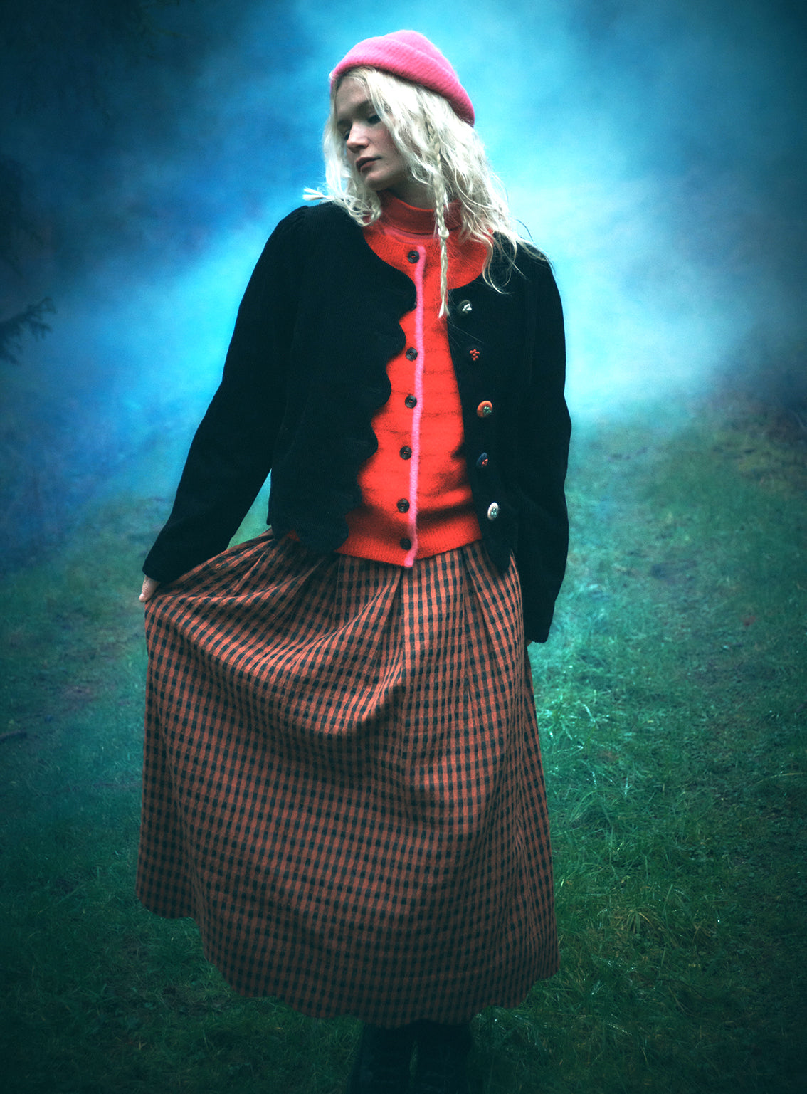 A woman stands outdoors in a misty blue-green environment wearing a bright pink beret, a black cardigan over a vivid orange-red top, and a brown-and-black checkered skirt.