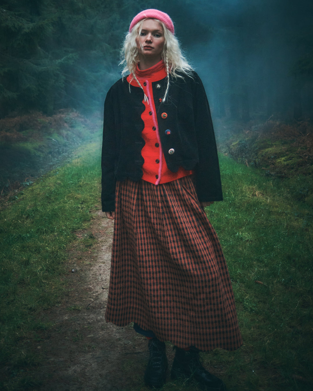 A model stands on a misty forest path wearing a pink beret, a red ruffled cardigan over a black jacket, and a red and black checkered skirt.