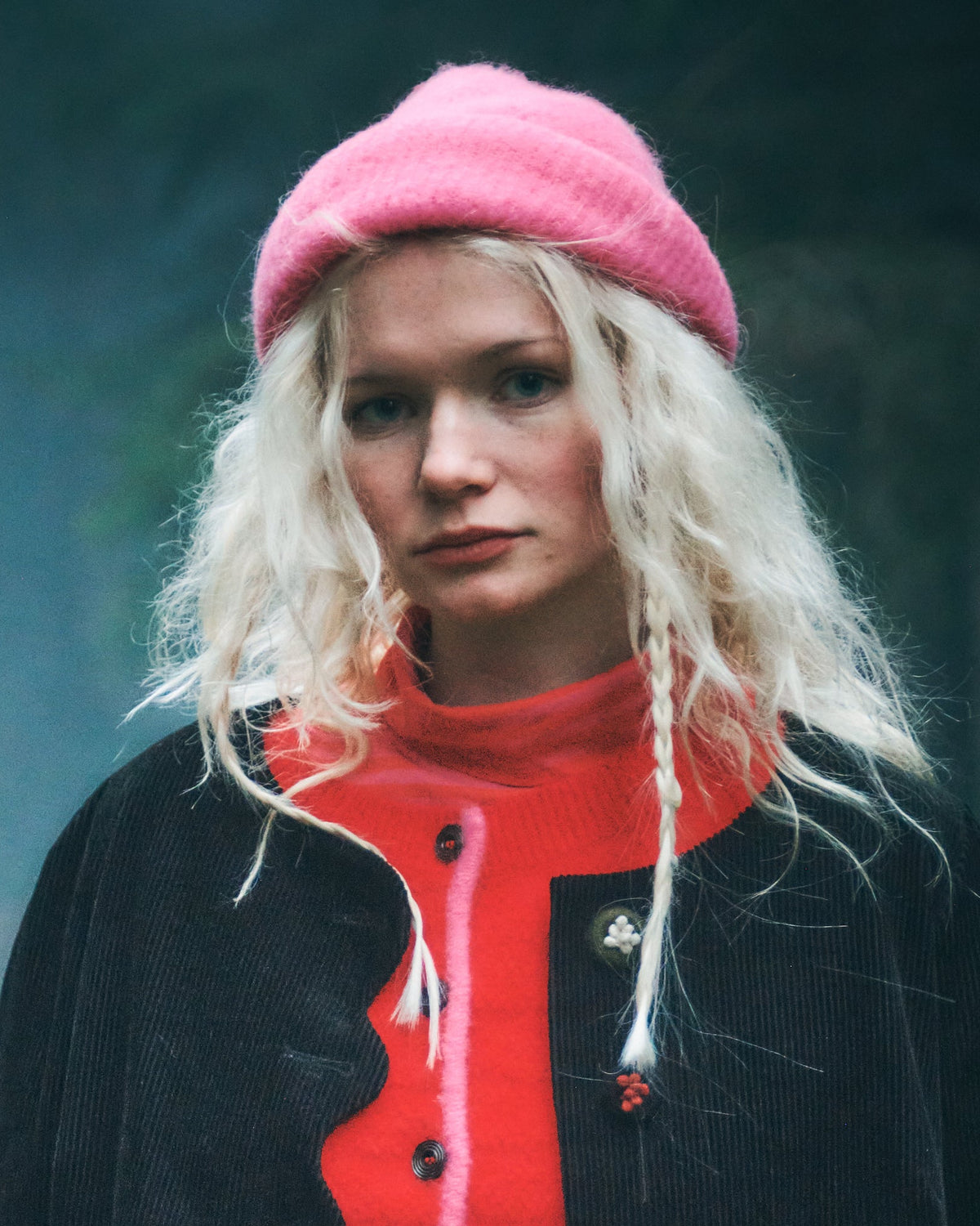 A woman with long blonde hair wearing a pink knit beret, a bright red scalloped sweater under a black jacket, and a brown plaid skirt, standing in a dark, misty outdoor setting.