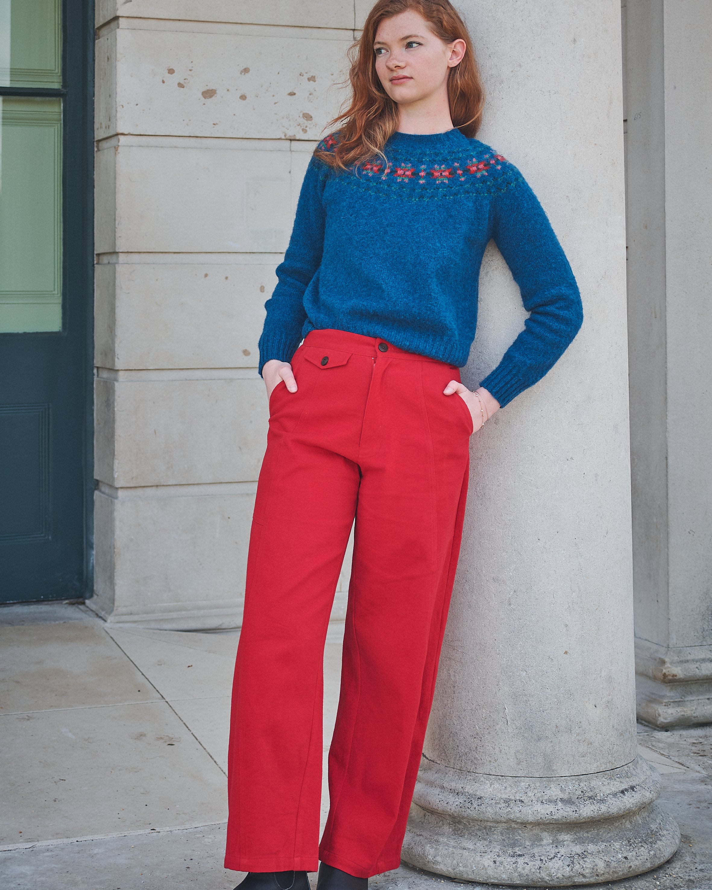 Woman wearing a blue sweater with red patterns and red pants standing against a stone building.