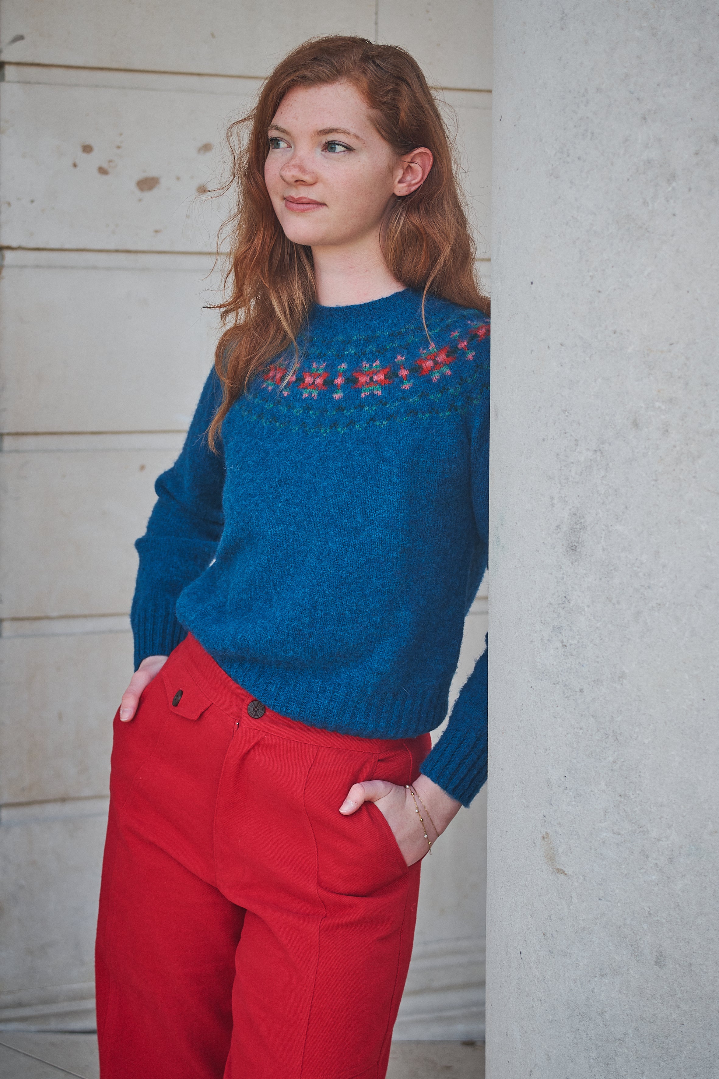 Woman wearing a blue sweater with red patterns and red pants against a light-colored wall.
