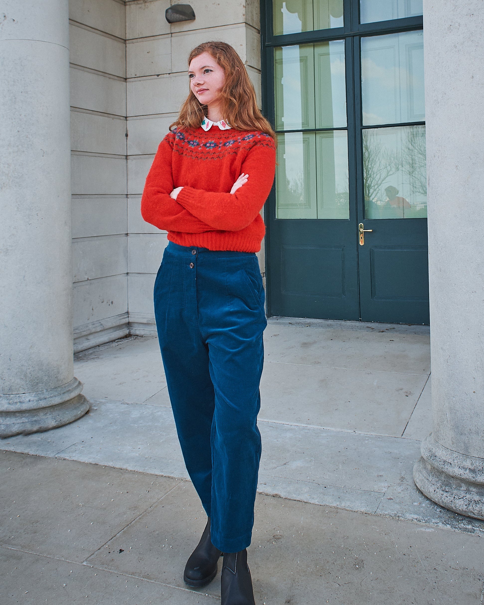Person wearing a red sweater and blue pants standing in front of stone columns and a window.