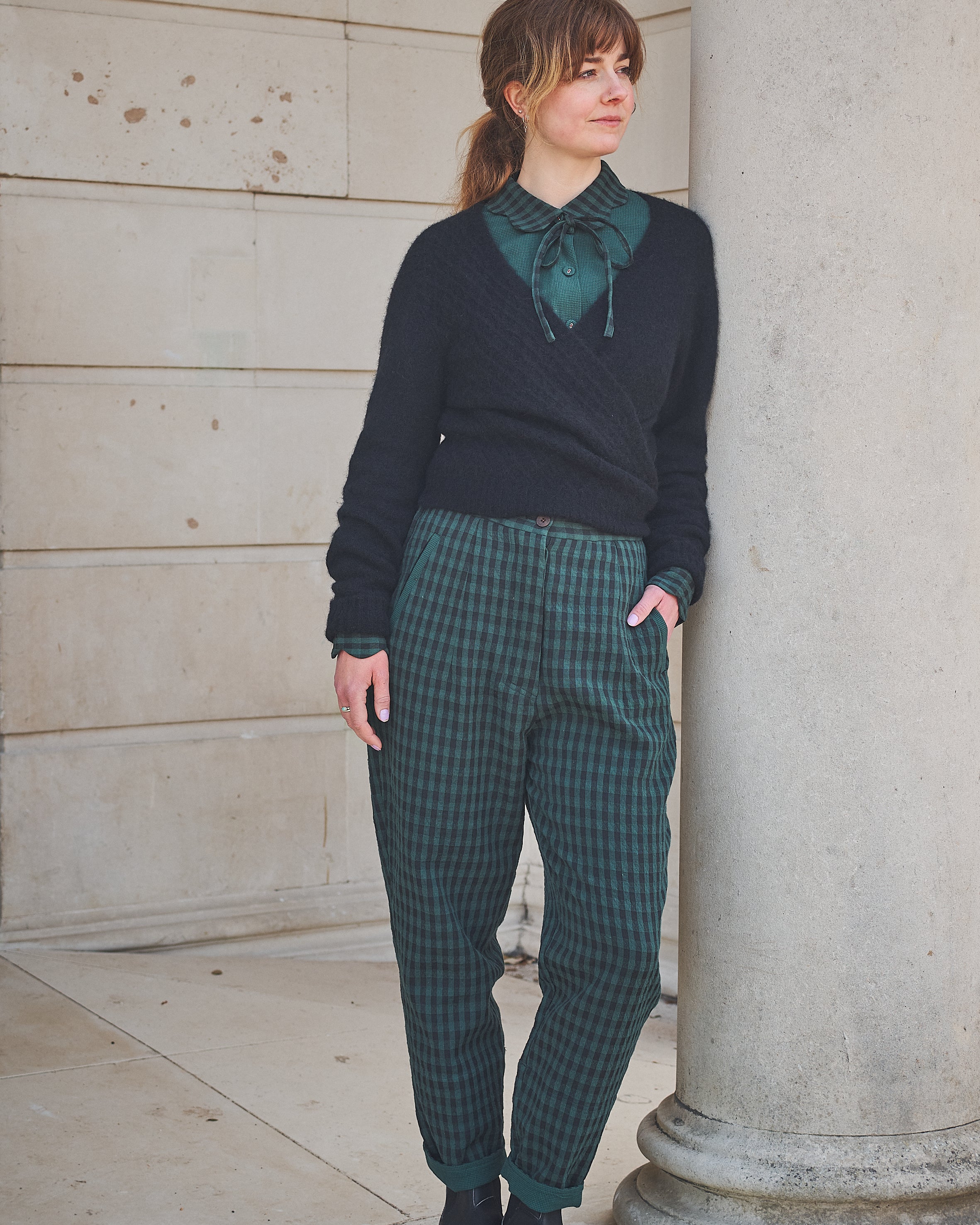 Woman wearing a dark sweater and green plaid pants standing against a stone wall.