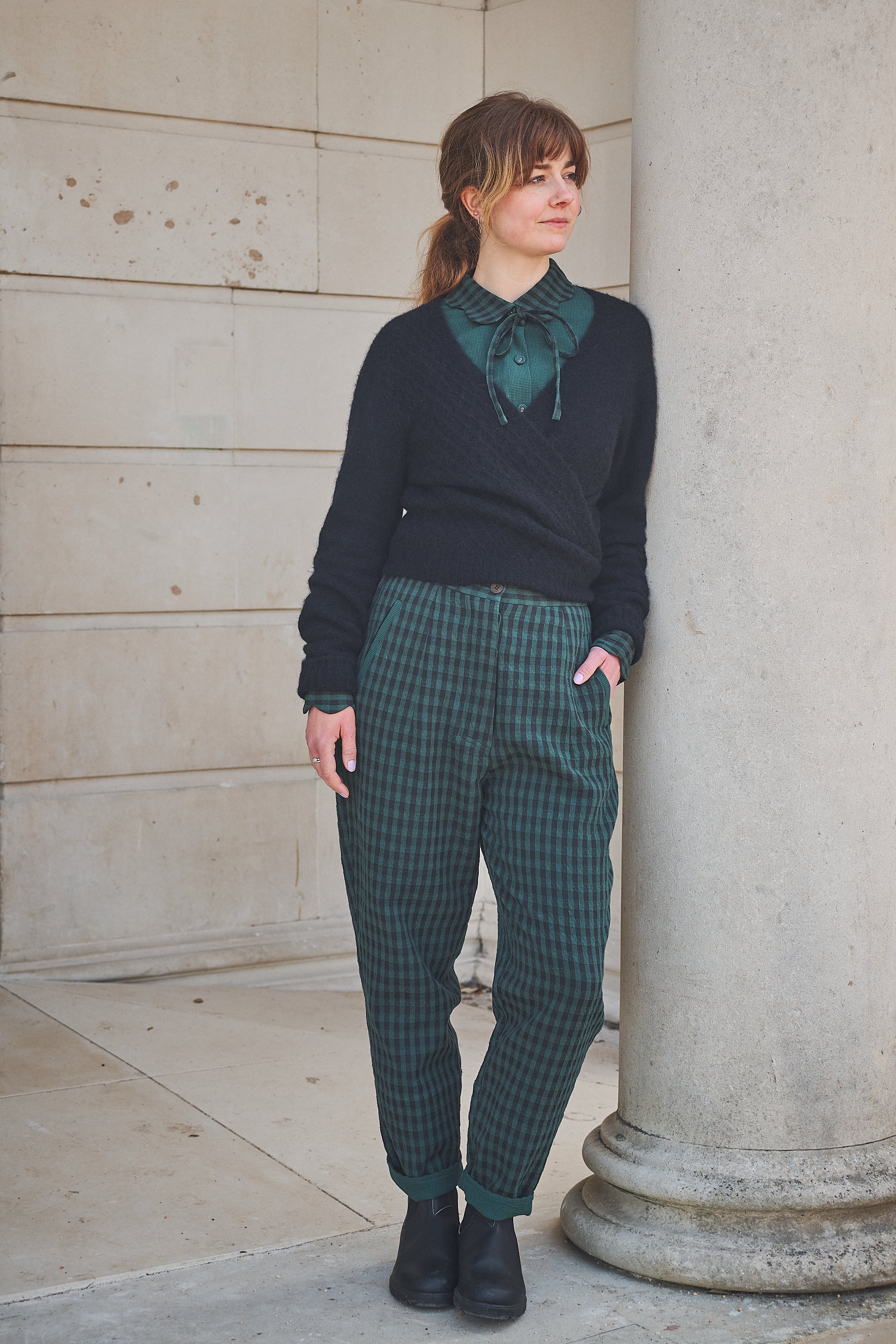 Woman wearing a dark sweater and green plaid pants standing against a stone wall.