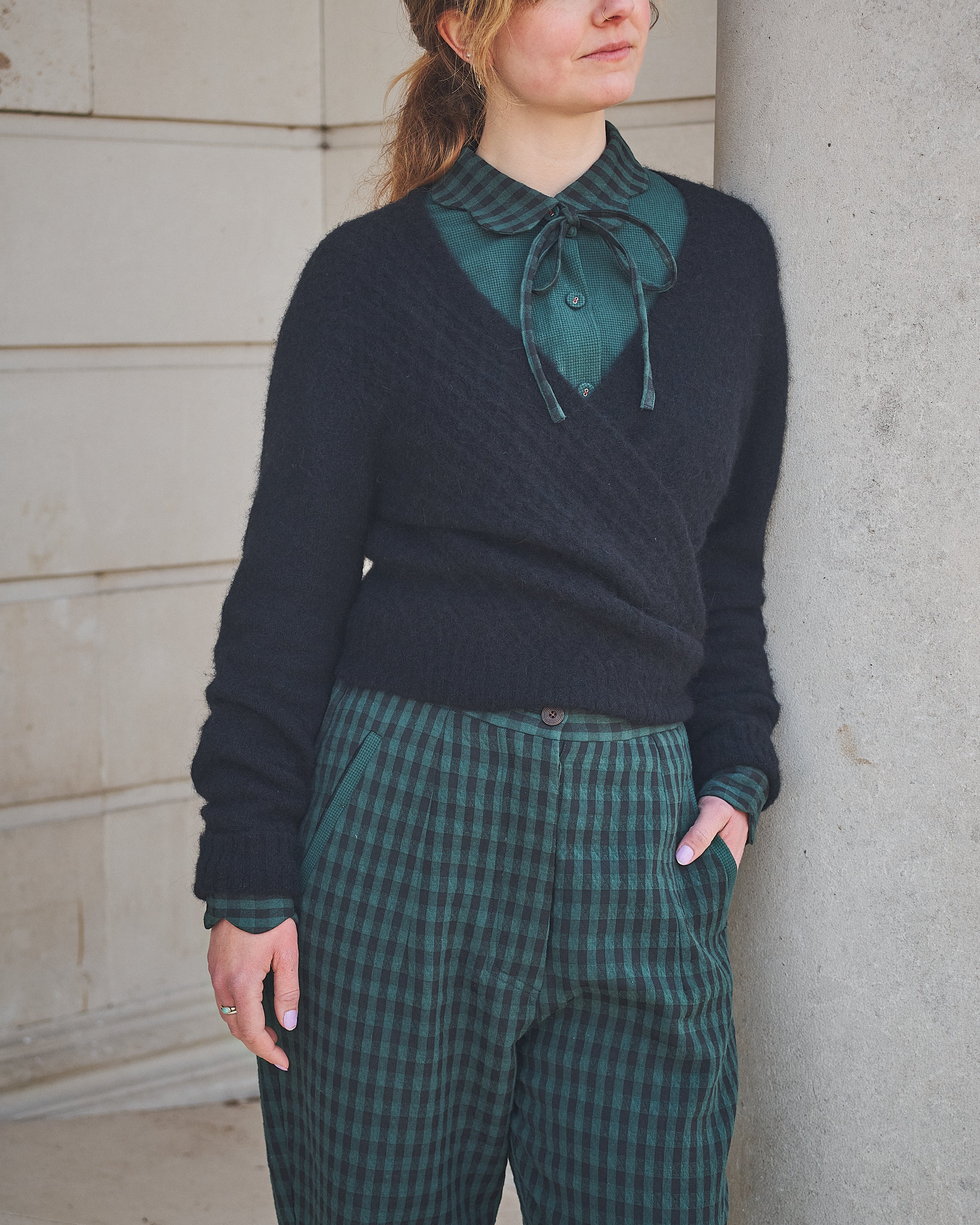 Woman wearing a dark sweater over a green shirt with plaid pants, standing against a concrete wall.