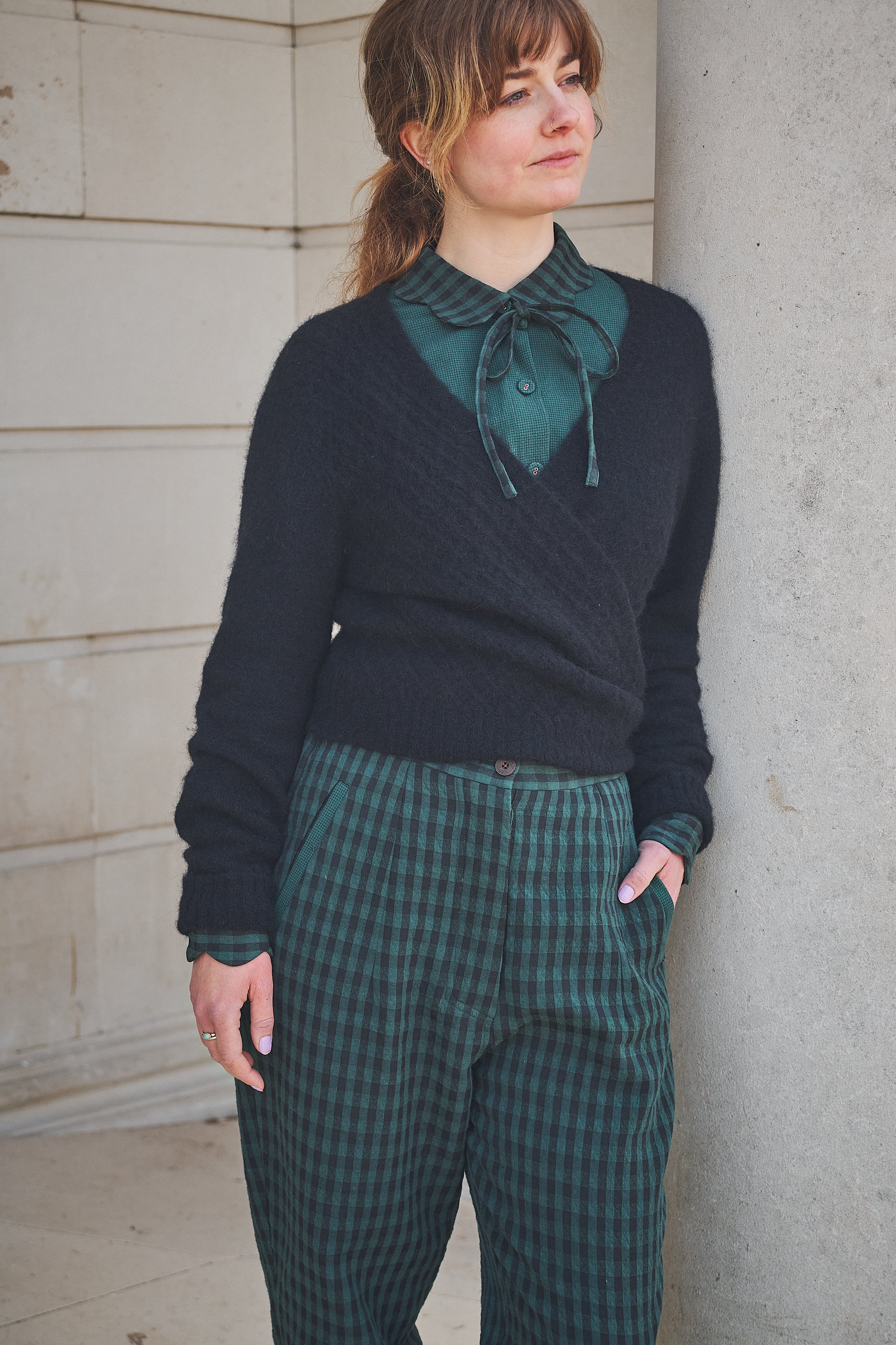 Woman wearing a dark sweater over a green shirt with plaid pants, standing against a concrete wall.