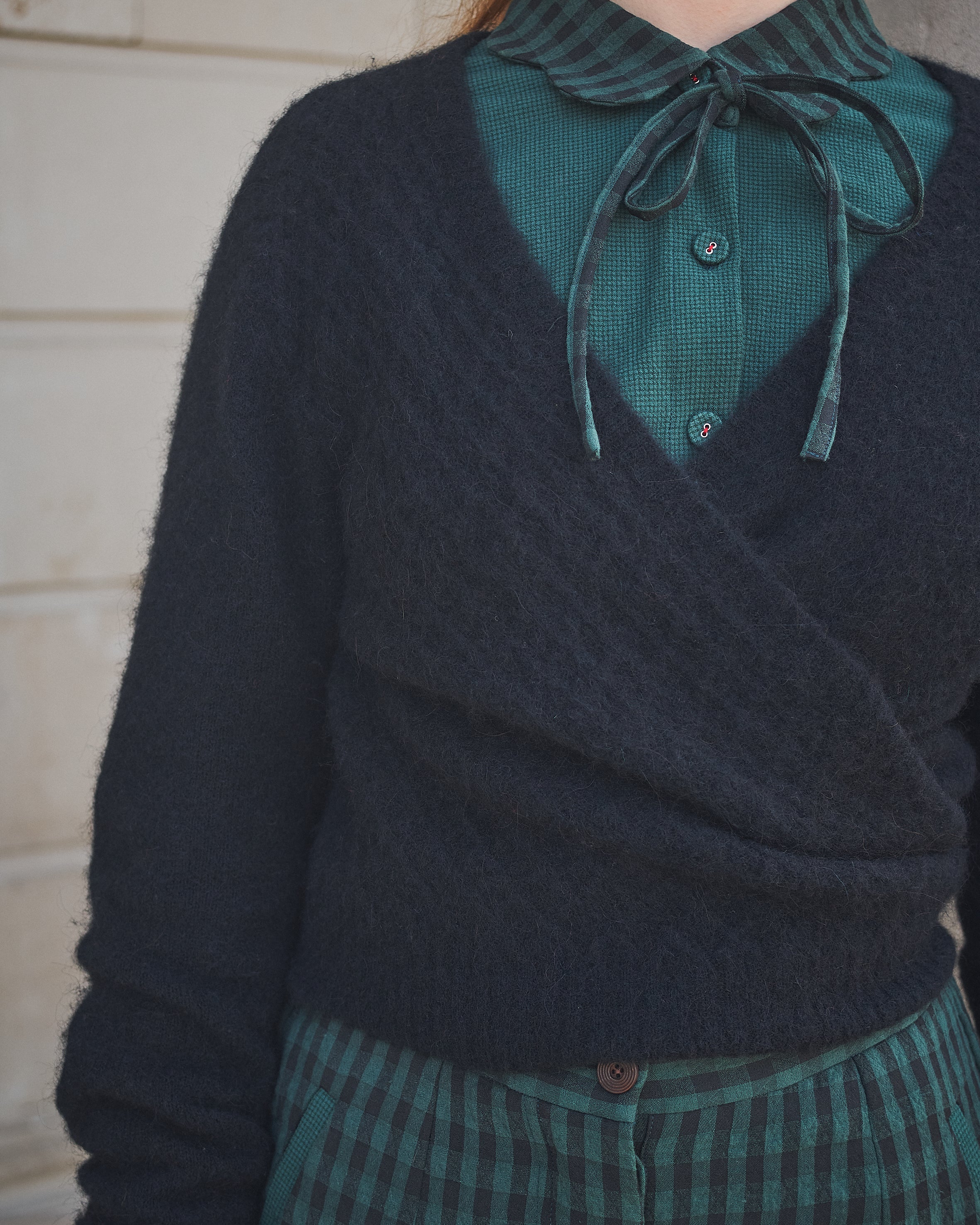 Person wearing a dark sweater over a green shirt with a bow tie, standing against a neutral background.