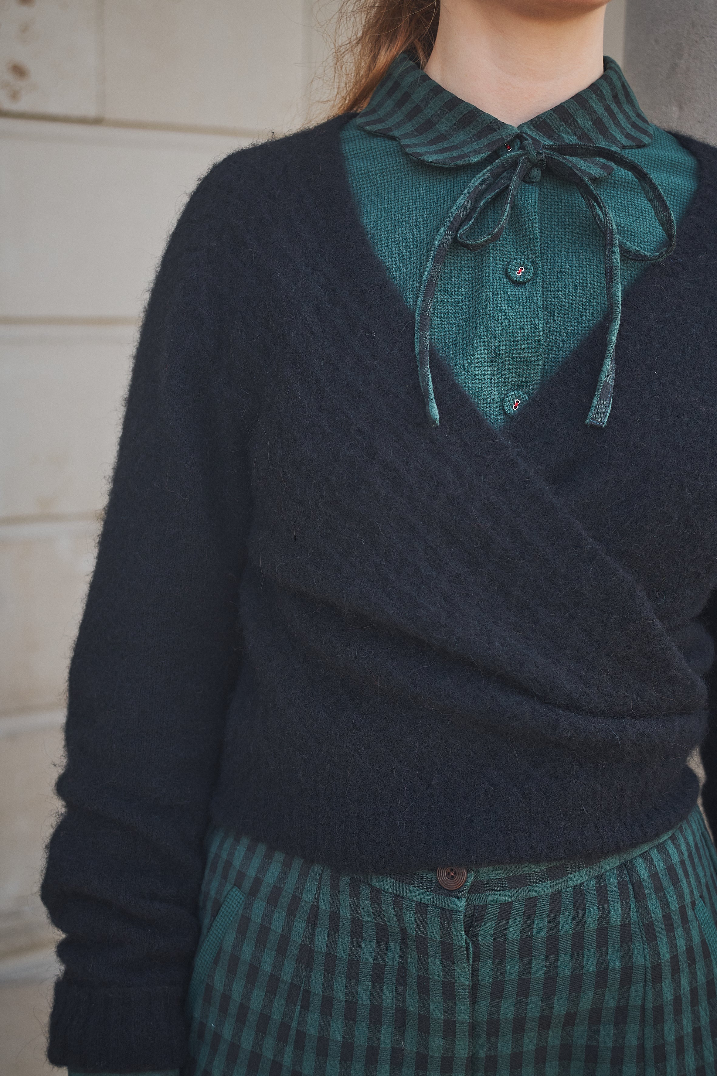 Person wearing a dark sweater over a green shirt with a bow tie, standing against a neutral background.