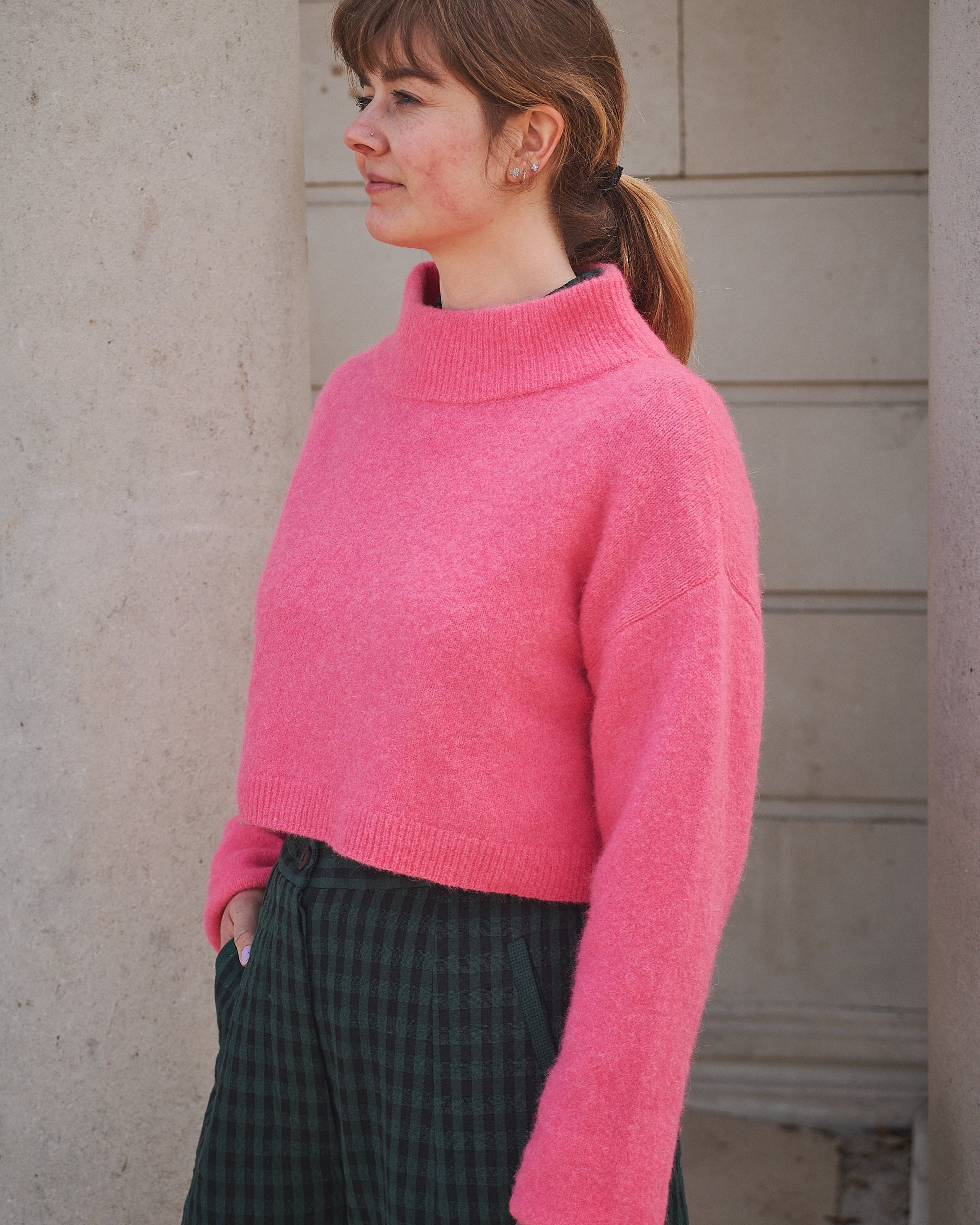 Person wearing a pink sweater and plaid pants standing against a neutral wall.