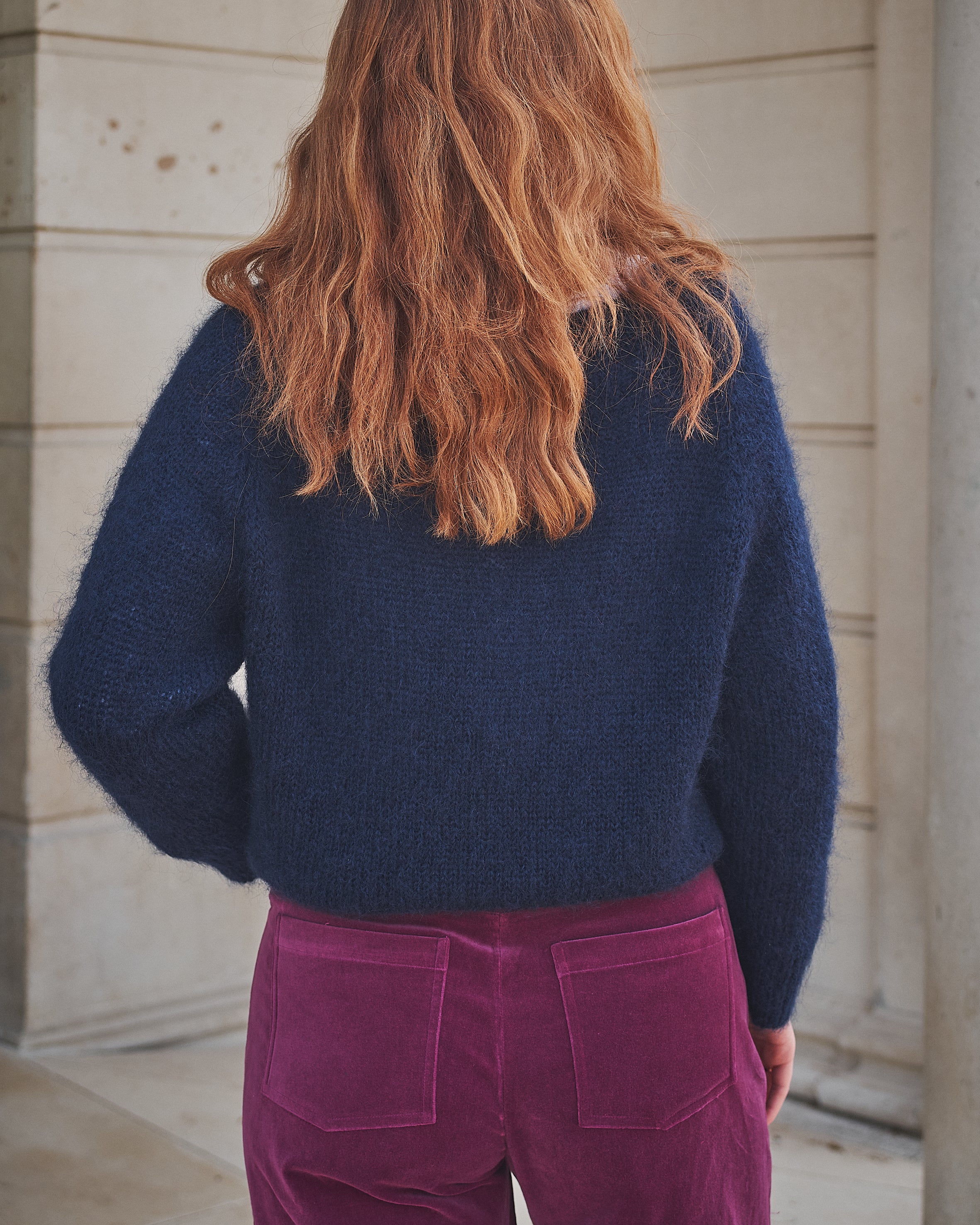 Person wearing a navy sweater and pink pants standing against a stone wall.