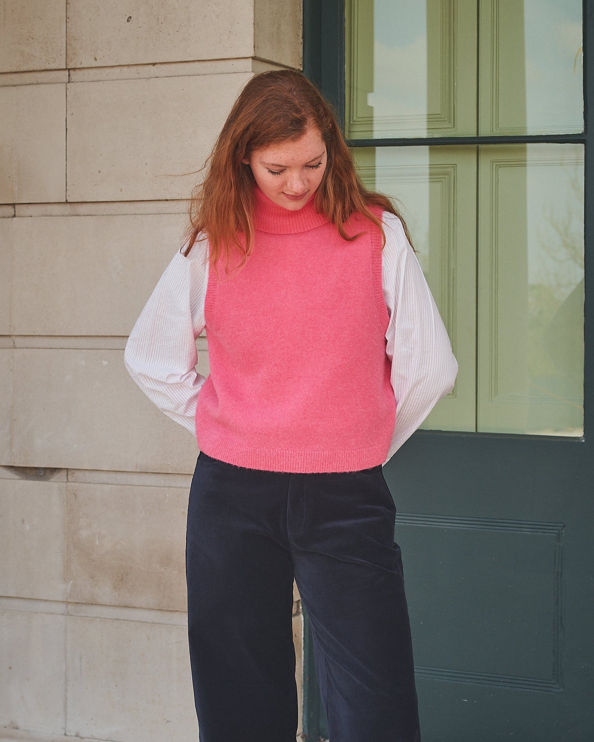 Person wearing a pink vest over a white shirt with dark pants, standing against a building.