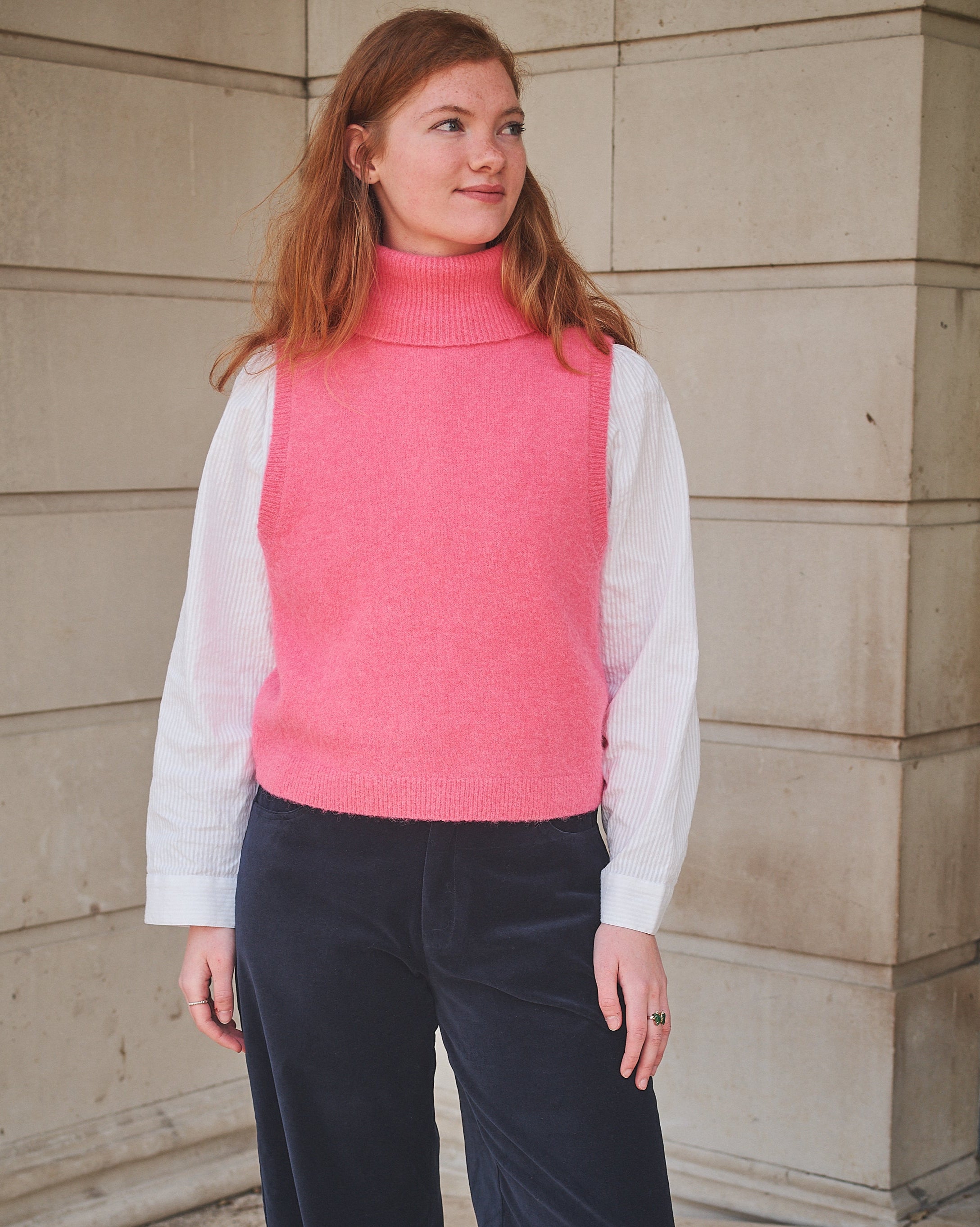 Woman wearing a pink vest over a white shirt and dark pants against a beige wall.