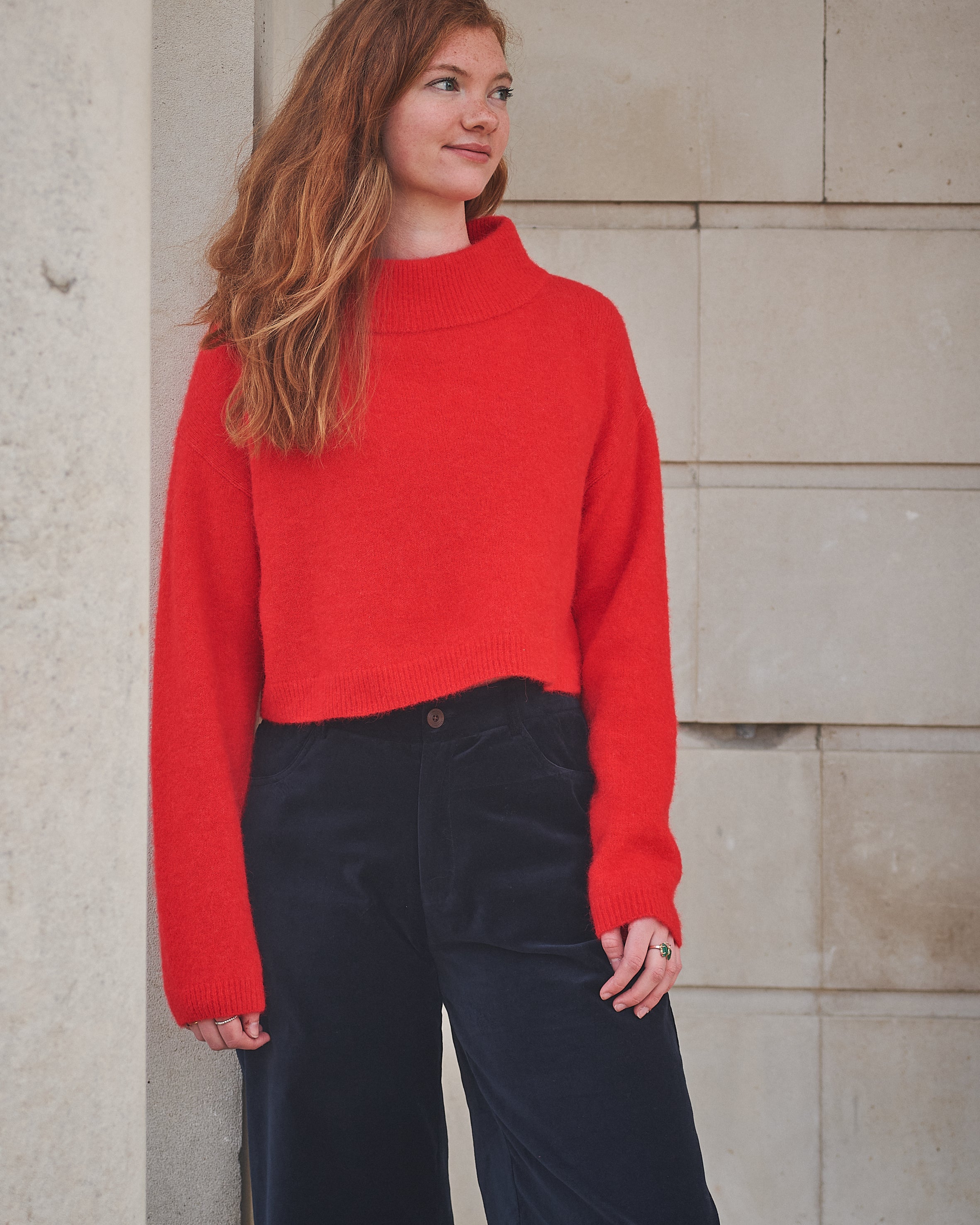 Woman wearing a red sweater and dark pants standing against a stone wall.