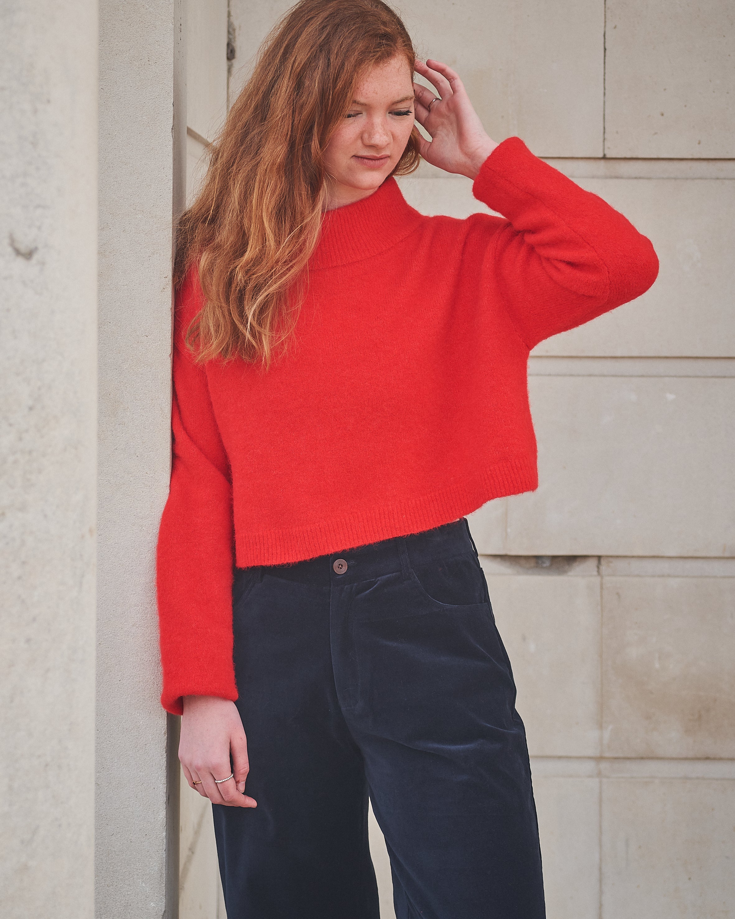 Woman wearing a red sweater and dark pants against a stone wall.