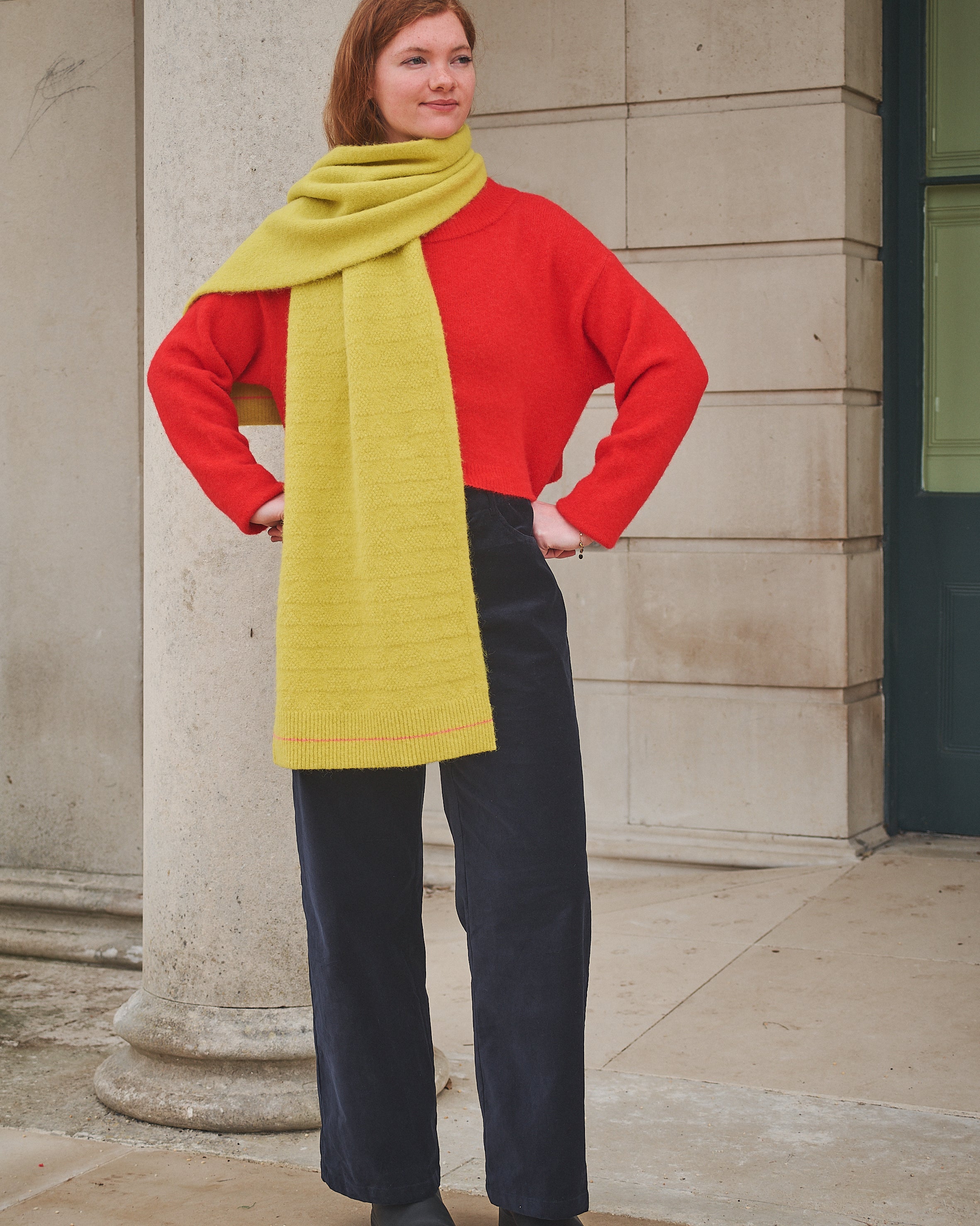 Woman wearing a red sweater, yellow scarf, and black pants standing against a stone building.
