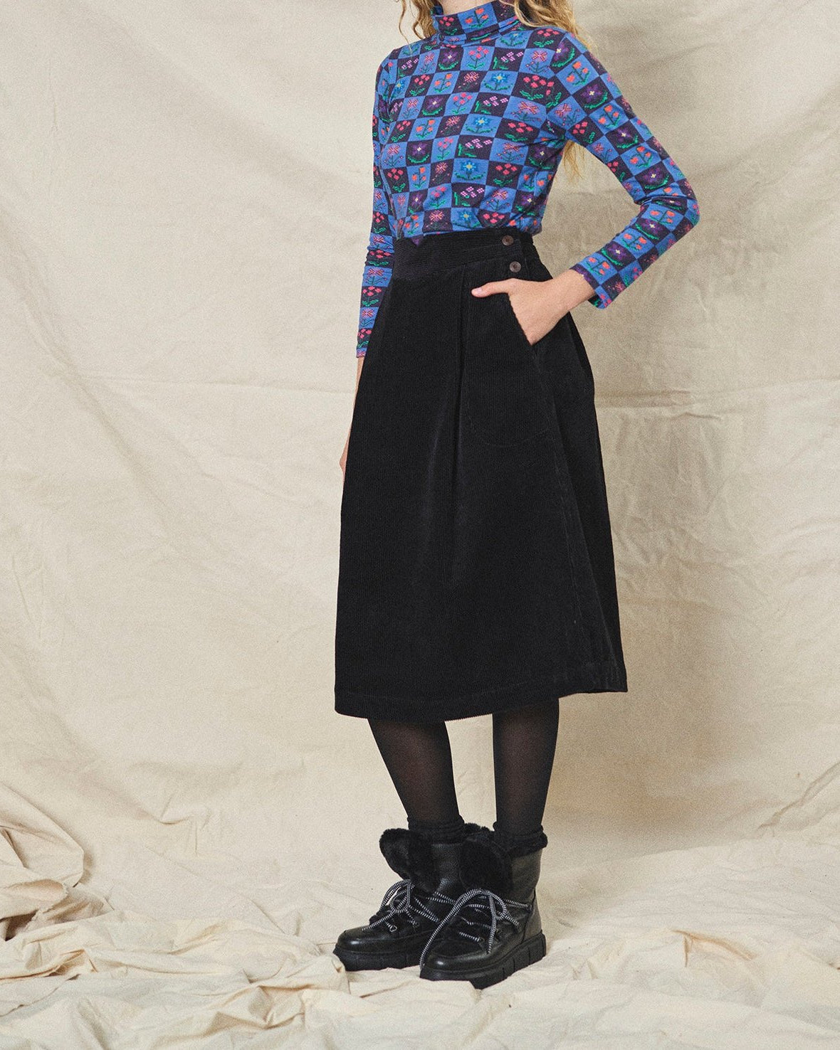 A person wearing a straight black corduroy skirt with a shaped waistband and deep inverted pleats, paired with a multicolored floral top and black boots.