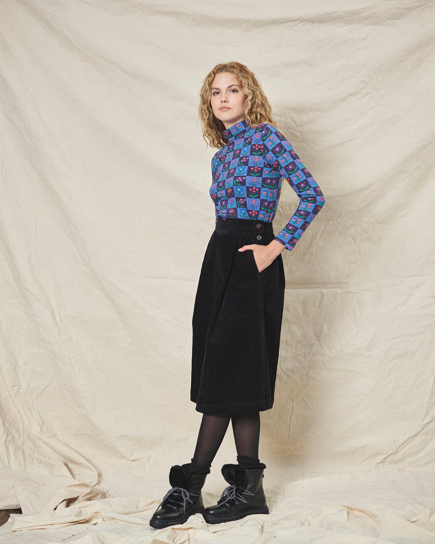 A person wearing a straight black corduroy skirt with a shaped waistband and deep inverted pleats, paired with a multicolored floral top and black boots.