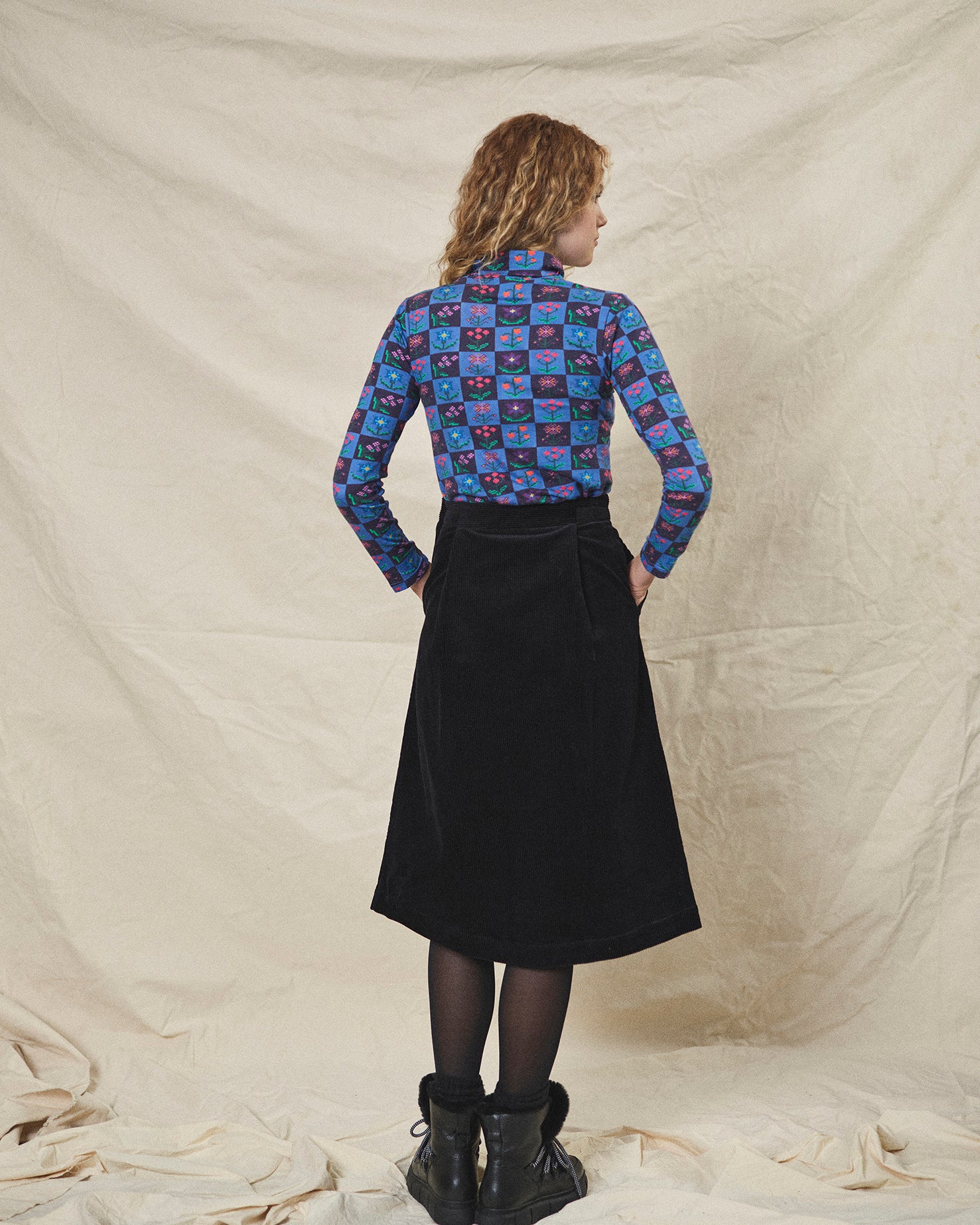 A person wearing a straight black corduroy skirt with a shaped waistband and deep inverted pleats, paired with a multicolored floral top and black boots.